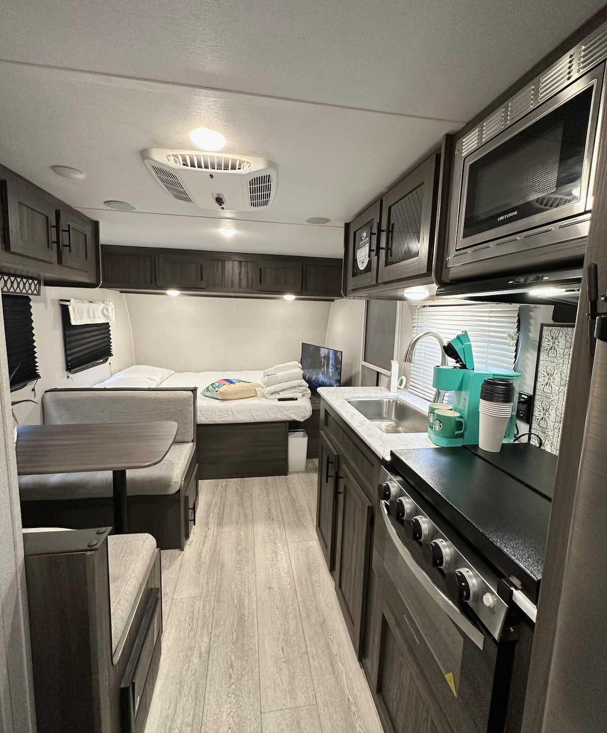 The modern camper interior features a functional kitchen with a microwave, sink, and coffee station. A booth dinette is present, with seating for four and a potential sleeping space. A Queen bed is positioned in the rear, accompanied by overhead cabinets and ambient lighting.
