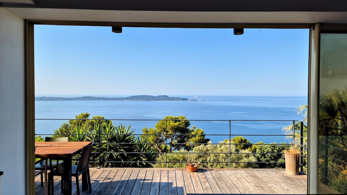 A wide terrace opens up to a panoramic view of the sea, with islands visible in the distance. A wooden dining table is situated on the terrace, surrounded by greenery, while large glass doors frame the scene, allowing for an airy and bright atmosphere.