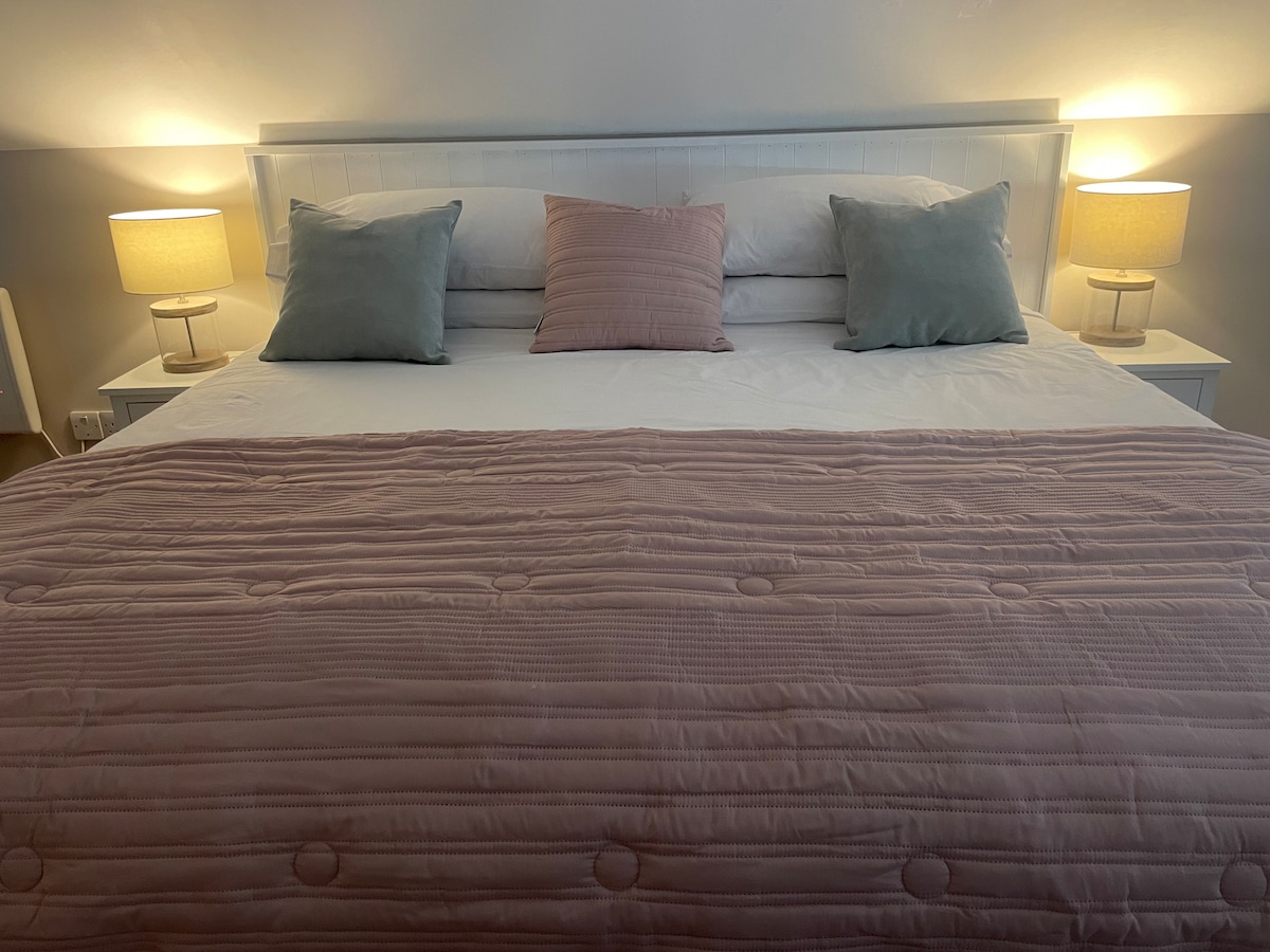 The sleeping area features a spacious bed with a soft pink quilted coverlet, complemented by two decorative pillows. Each side of the bed is flanked by warm table lamps, providing a gentle glow to the inviting atmosphere.