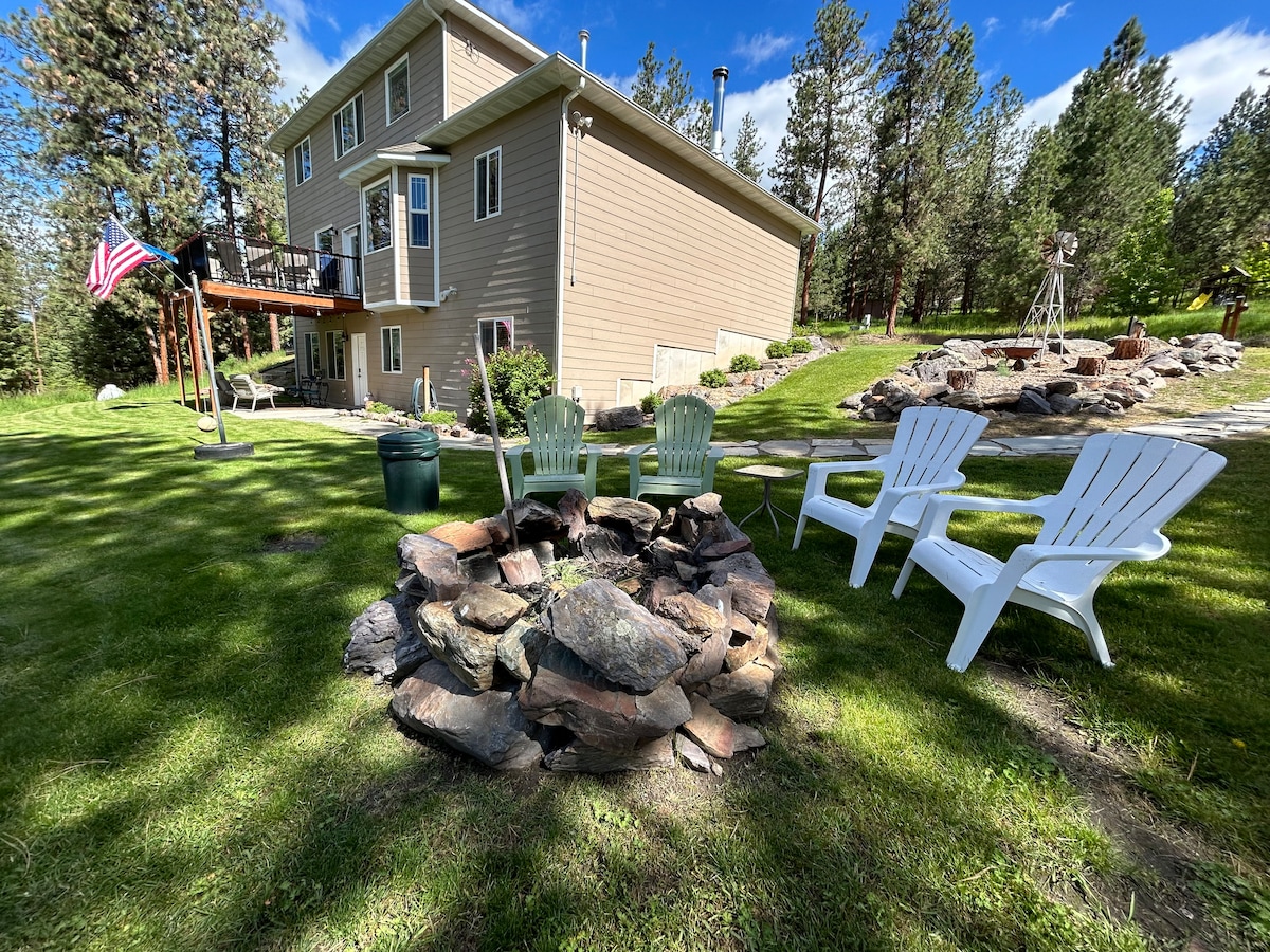 A fire pit made of natural stone is surrounded by several outdoor chairs, including two white adirondack chairs and two green chairs. The background features a modern two-story house and well-maintained greenery, with tall pine trees providing shade.