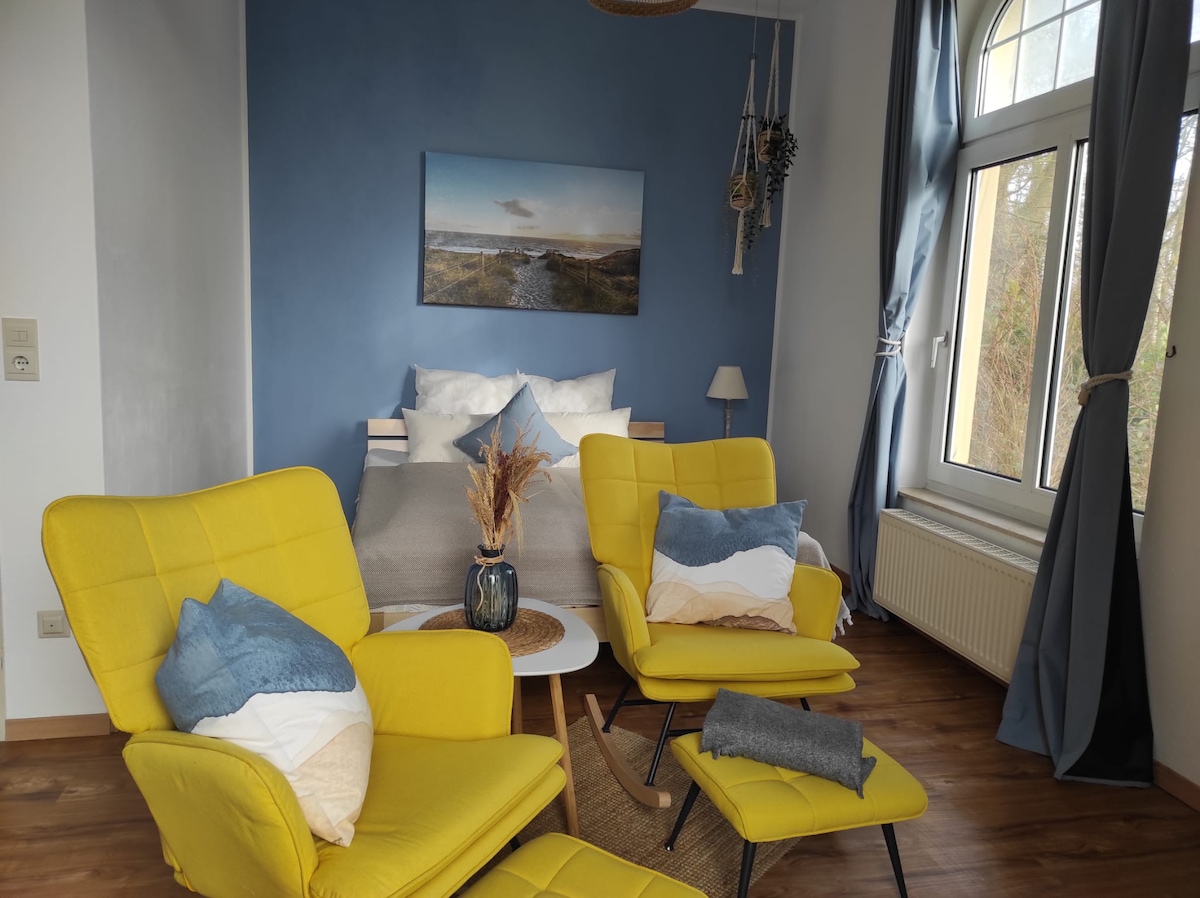 A bright living and sleeping area is displayed, featuring two yellow armchairs complemented by soft cushions. A small table holds a decorative vase, while a cozy double bed with crisp linens is positioned against a blue wall. Natural light streams through the large window, framed by long curtains.