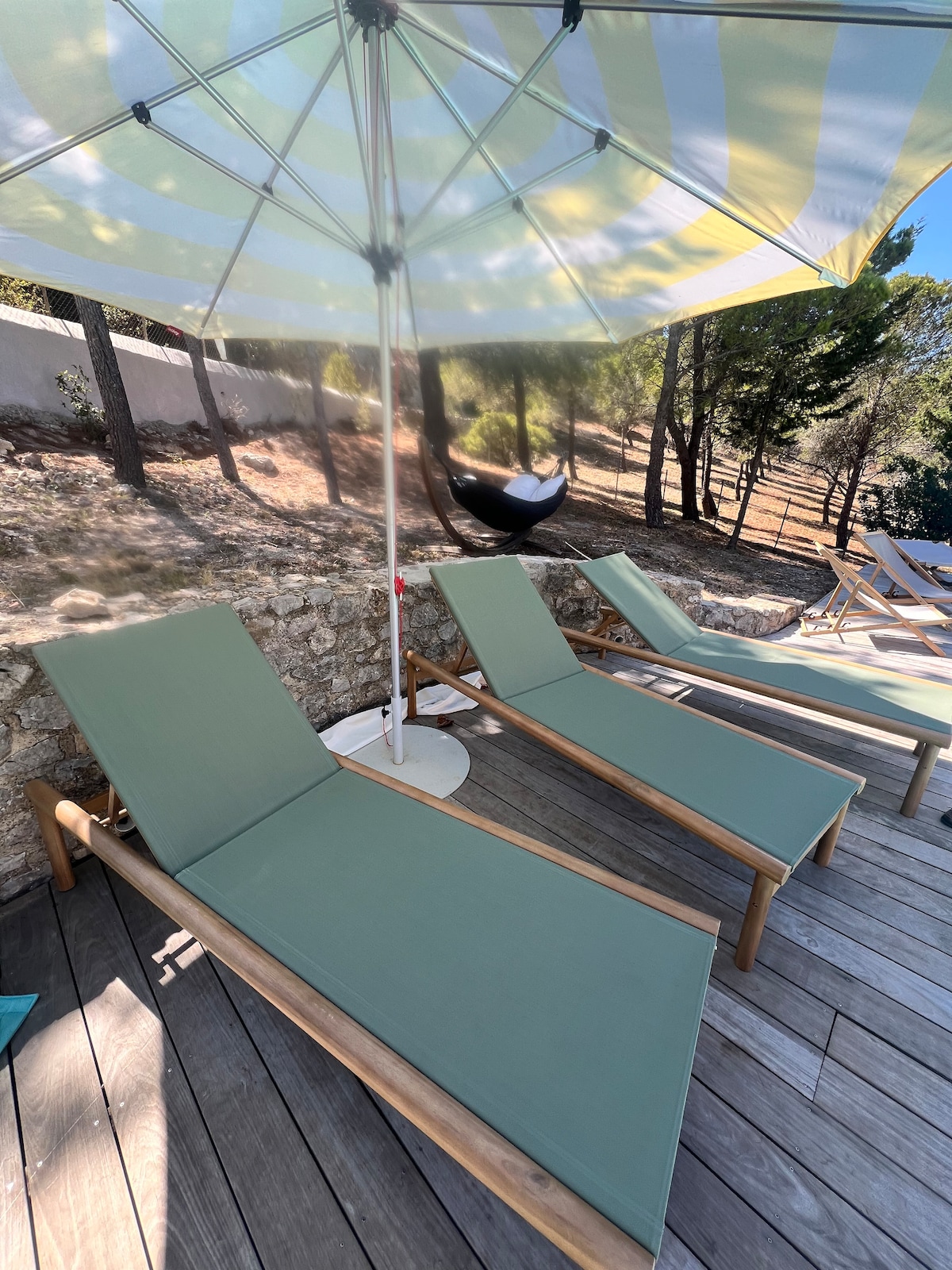 Two green sun loungers are positioned under a large striped umbrella, offering shade on a wooden deck. The area is surrounded by trees, providing a natural and serene setting. A stone wall is visible in the background, enhancing the tranquil atmosphere.