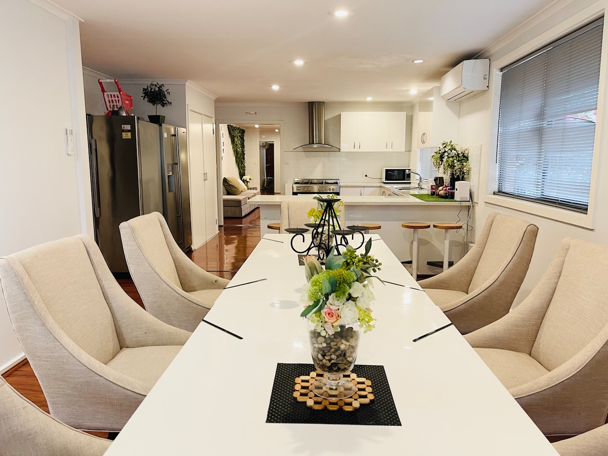 A modern dining area features a long white table surrounded by six soft, beige chairs. A decorative centerpiece with flowers adds a touch of elegance. The open kitchen in the background includes stainless steel appliances and plenty of natural light from the nearby window.