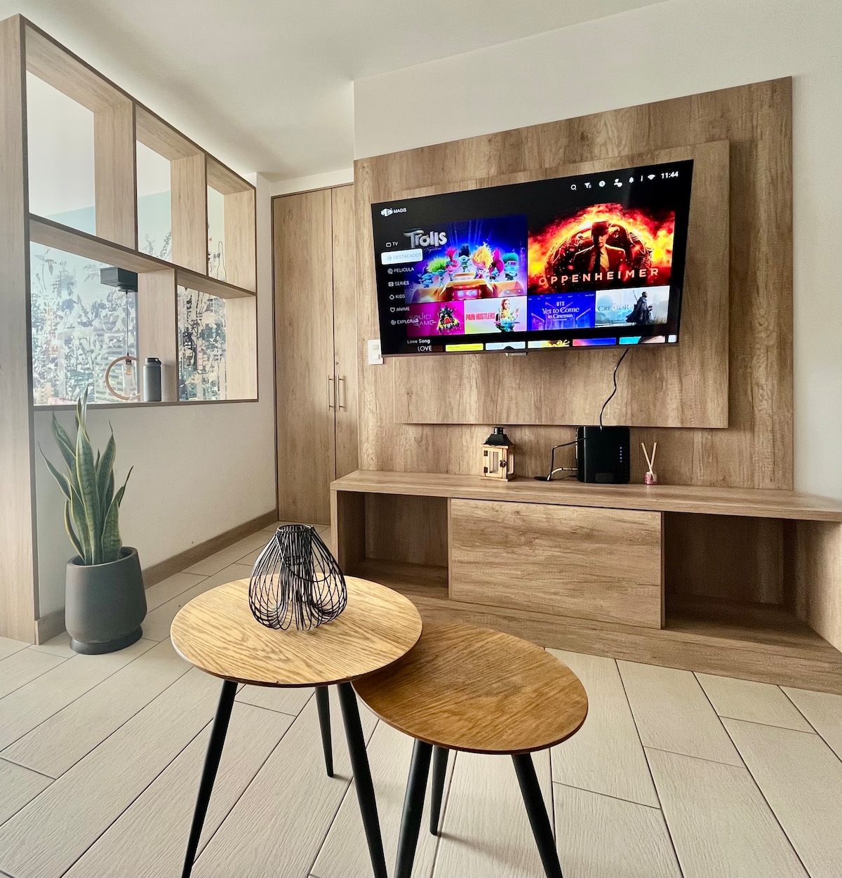 A modern living area features a mounted flat-screen TV displaying various applications. Two small wooden tables are positioned in front, alongside a decorative plant in a gray pot. Light-colored walls and wooden accents contribute to a clean, airy atmosphere.