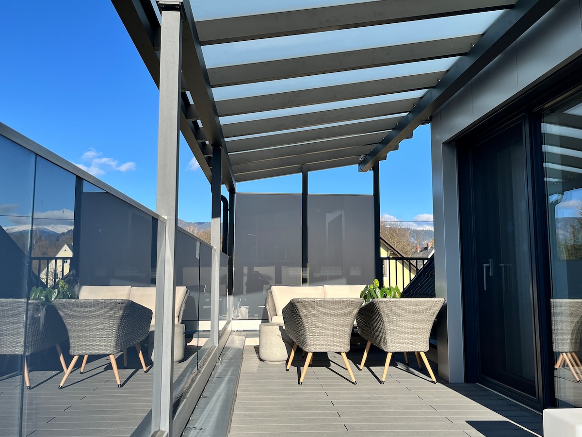 An outdoor terrace is enclosed by glass panels and features a pergola overhead. Two comfortable seating areas are arranged with woven chairs and a low table, complemented by greenery. Clear blue skies and distant mountains provide a scenic backdrop.