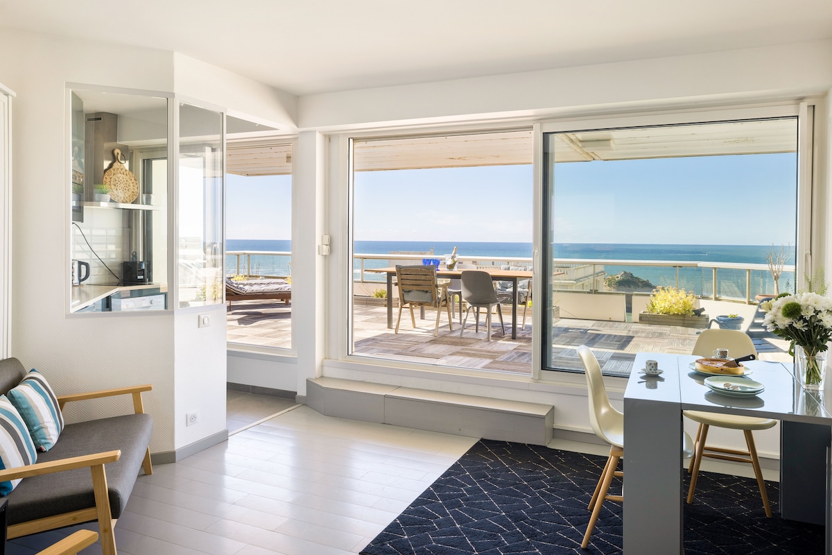 A bright living area features large glass windows providing unobstructed ocean views. A dining table is set nearby, complemented by a cozy sitting arrangement. The terrace can be seen through the glass doors, offering additional seating and space for outdoor enjoyment.