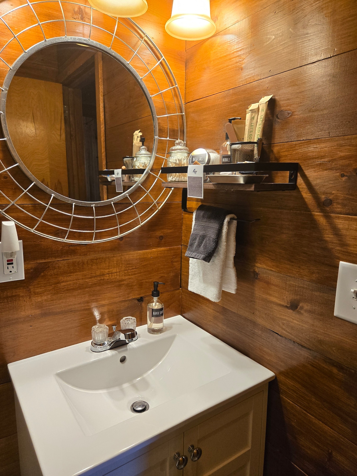 A compact bathroom features a white sink cabinet beneath a round mirror framed with metal. Wooden walls provide a warm ambiance. Shelves hold eco-friendly toiletries, and a hand towel is neatly arranged nearby. Soft lighting creates an inviting space.
