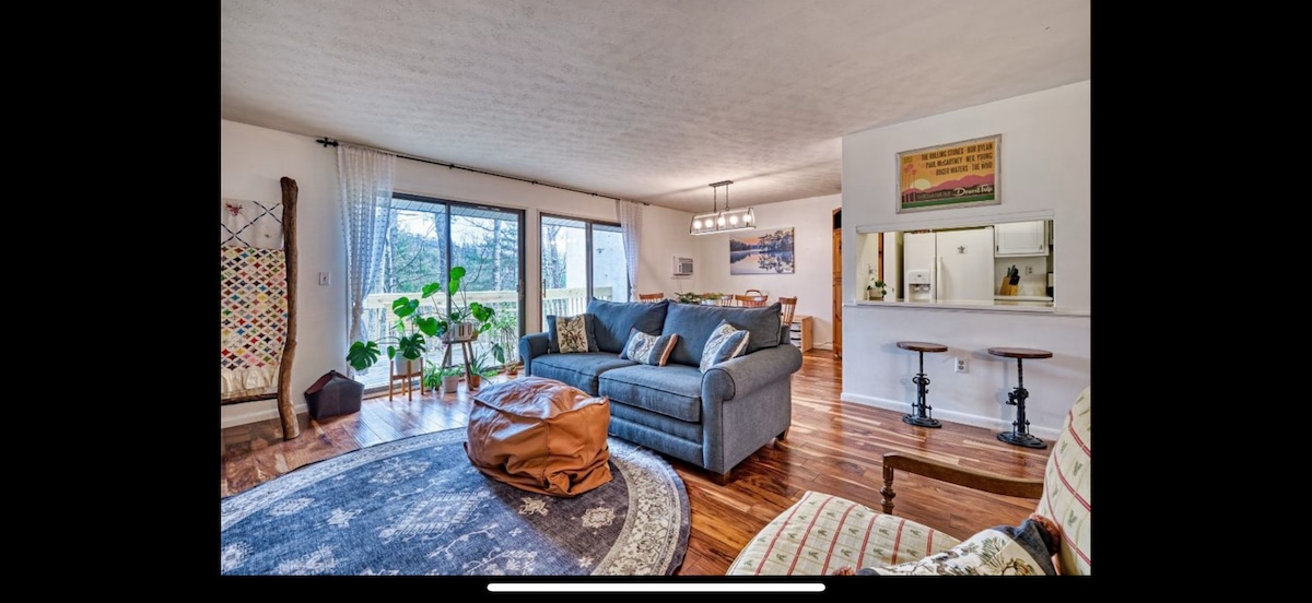 A spacious living area features a gray sofa adorned with decorative cushions, complemented by a brown pouf. Large windows allow natural light, revealing a view of greenery. A patterned area rug enhances the wooden flooring, while a dining area with a modern light fixture is visible in the background.