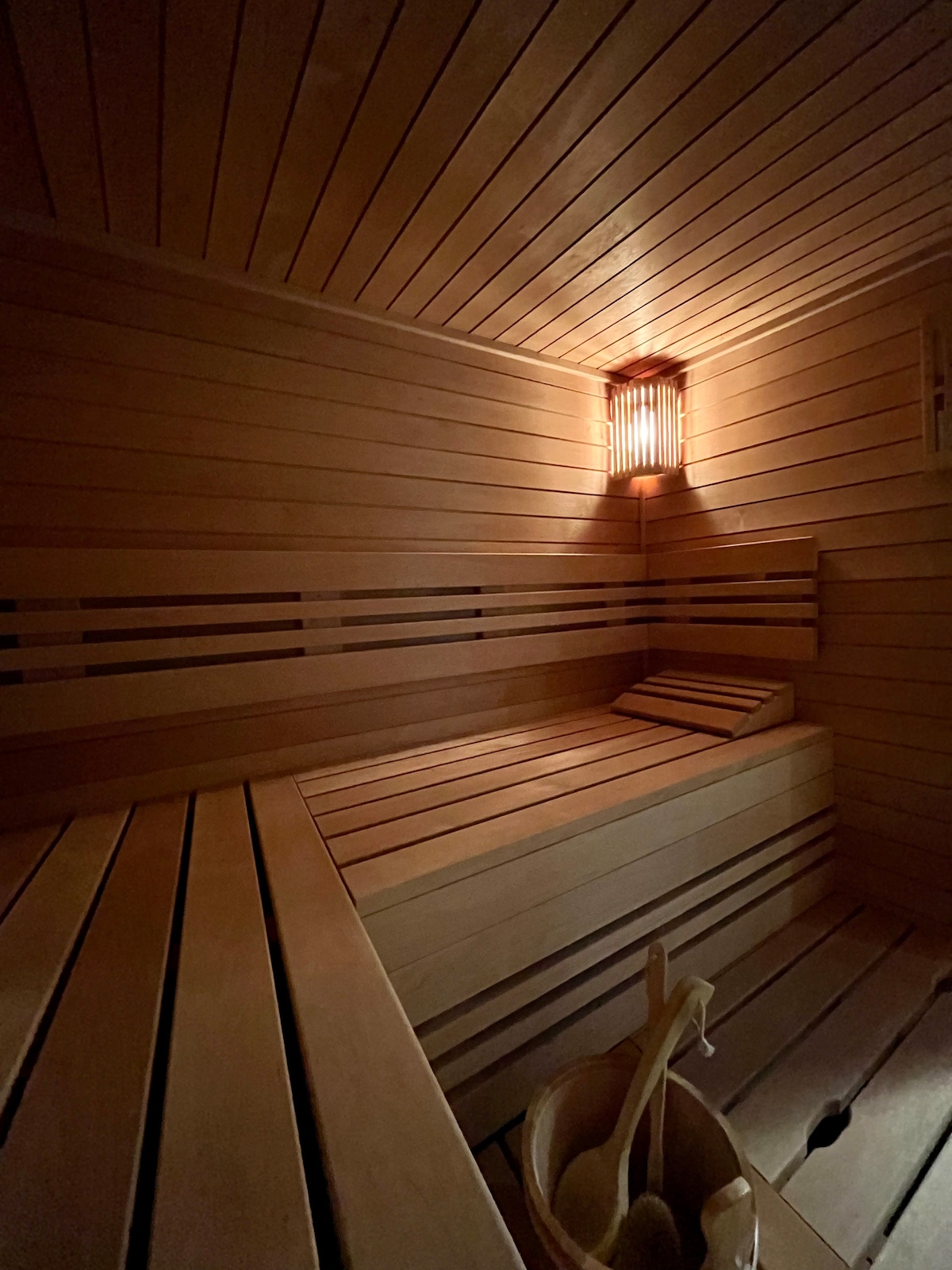 The wooden sauna features smooth, light-colored wooden panels along the walls and benches. Soft lighting emanates from a wall fixture, creating a warm ambiance. A small bucket sits beside the benches, complementing the serene environment designed for relaxation.