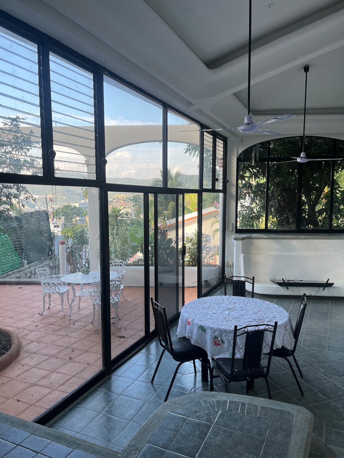 A bright dining area is depicted with a circular table surrounded by black metal chairs. Large glass panels provide views of a landscaped outdoor space. The room features a tiled floor and is complemented by earthy tones and natural light filtering through.