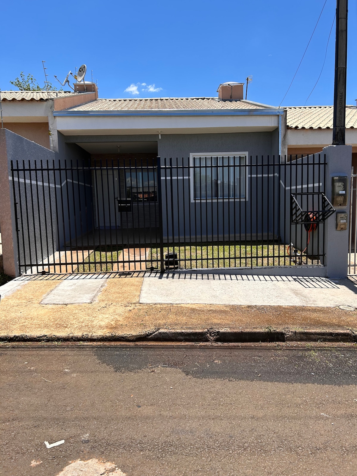 The exterior of a home is shown, featuring a grey façade and a modern black wrought-iron gate. A small patch of grass is visible in front of the house, with concrete pathways leading to the entrance. The sky is clear and blue.