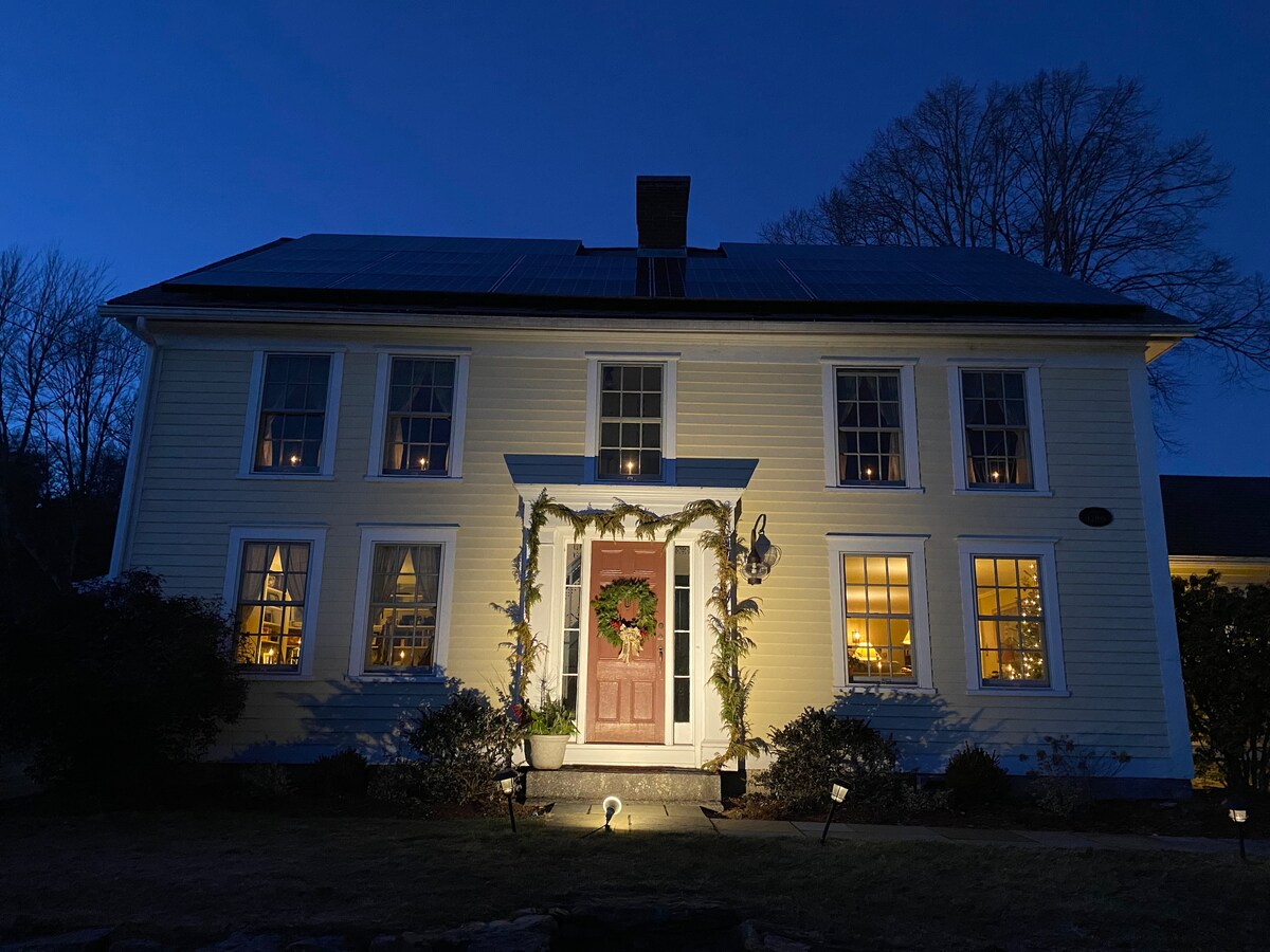 A stately farmhouse stands illuminated against the night sky. Warm light glows from the windows, highlighting the inviting entrance adorned with a wreath and seasonal decor. The exterior features classic architectural elements, including multiple windows and a welcoming front door.