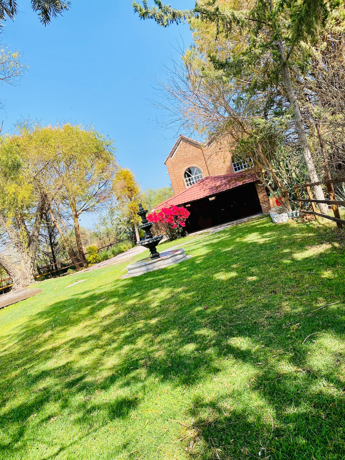 A spacious green lawn is featured, surrounded by trees and a pathways. A two-story brick house with a maroon roof stands under clear blue skies. A decorative fountain is positioned on the grass, complemented by vibrant pink flowers nearby.