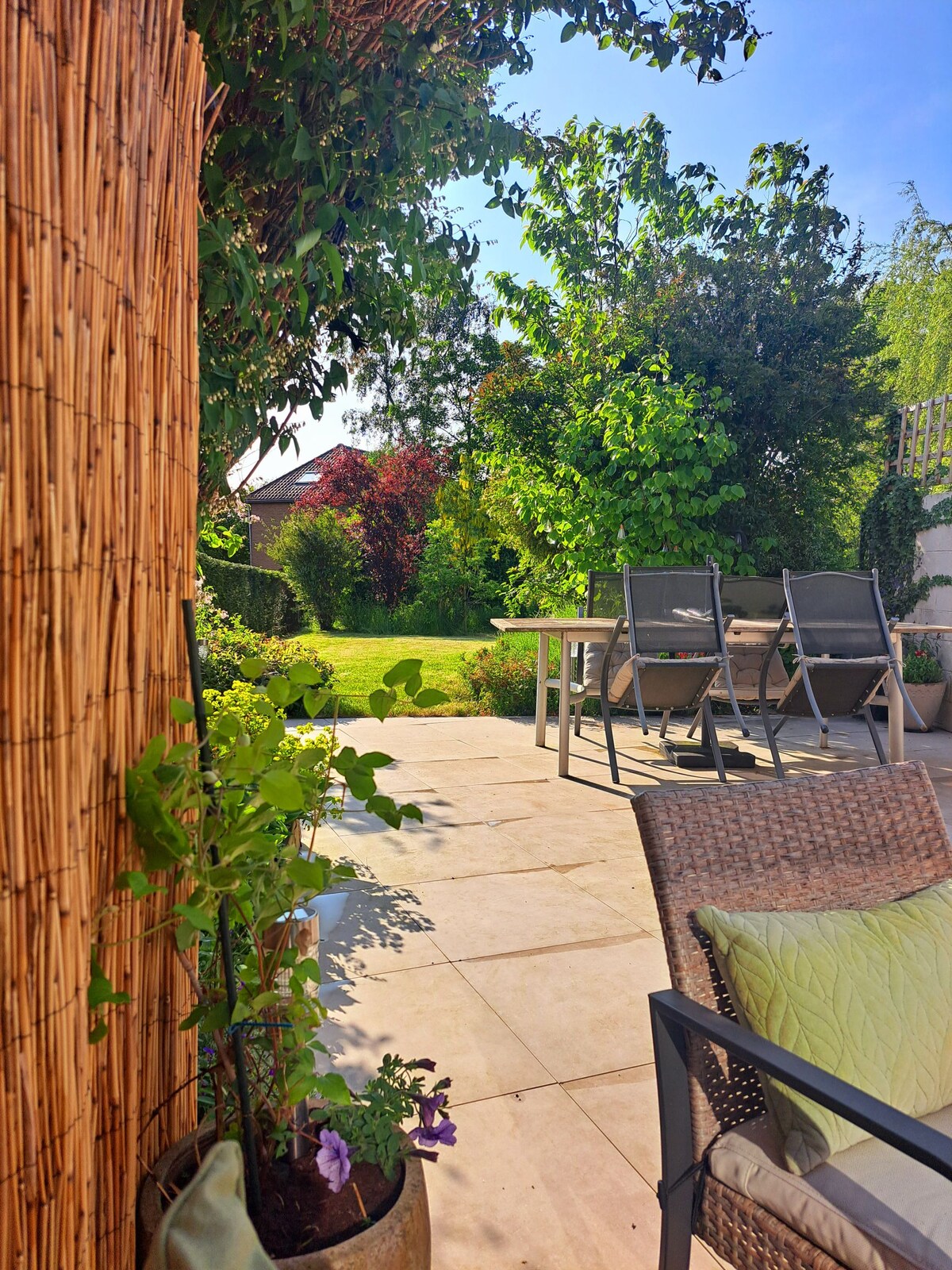 A private outdoor space is visible, featuring a large grassy area surrounded by trees and shrubs. A table with several chairs is positioned on a tiled patio. Potted plants add a touch of greenery, enhancing the serene environment.