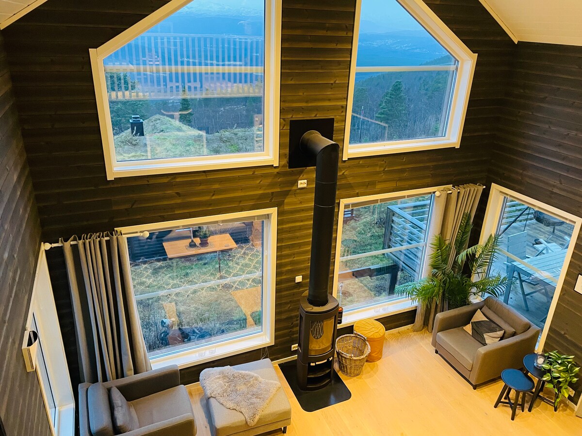 A spacious living area is presented with large windows offering expansive views of the surrounding landscape. A modern wood-burning stove is centrally located, complemented by two gray sofas and a small side table. Decorative plants add a touch of greenery to the inviting space.