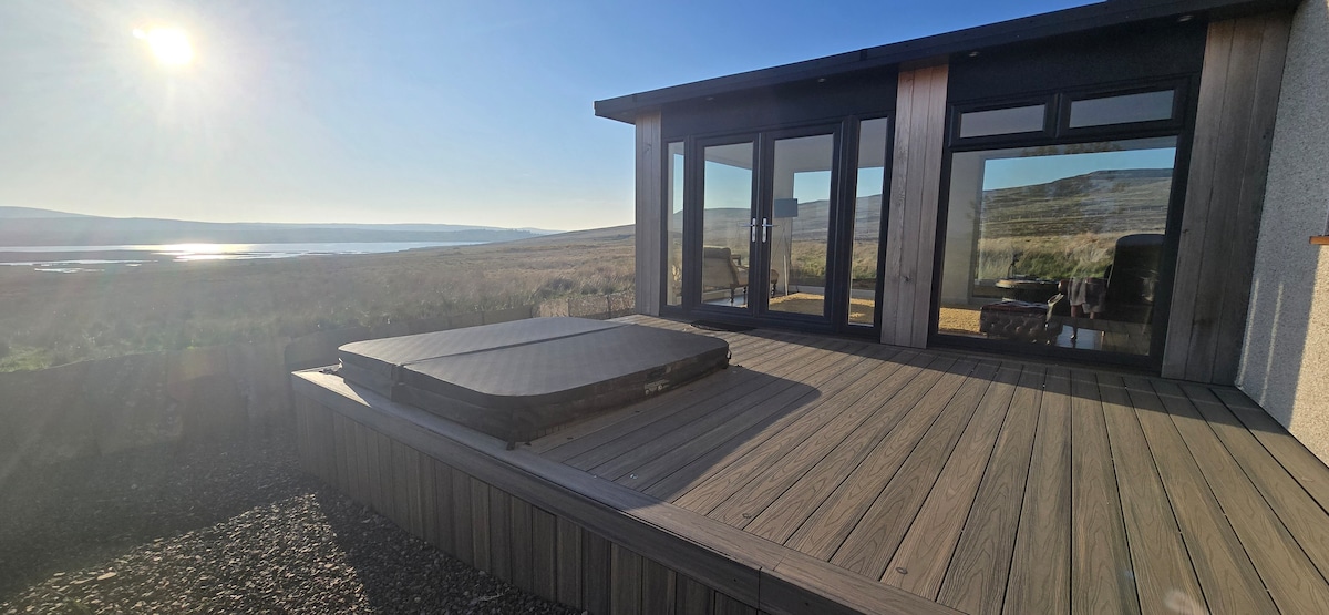 A spacious deck features a hot tub positioned for panoramic views of the surrounding landscape. Large glass doors provide a connection to the interior space, while sunlight illuminates the area, highlighting the natural beauty of the environment.