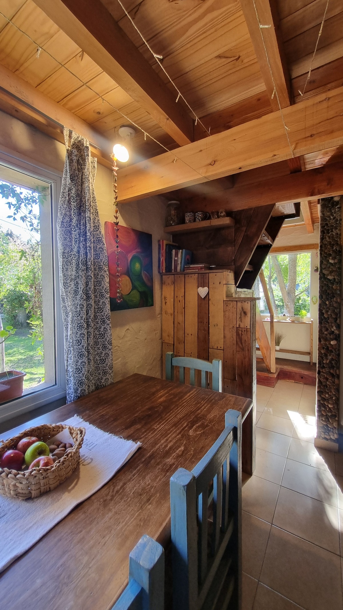 A cozy dining area is featured with a wooden table, surrounded by blue chairs. Sunlight illuminates the space through a window, highlighting a basket of apples on the table. Wooden beams are visible on the ceiling, and artwork is displayed on the wall.