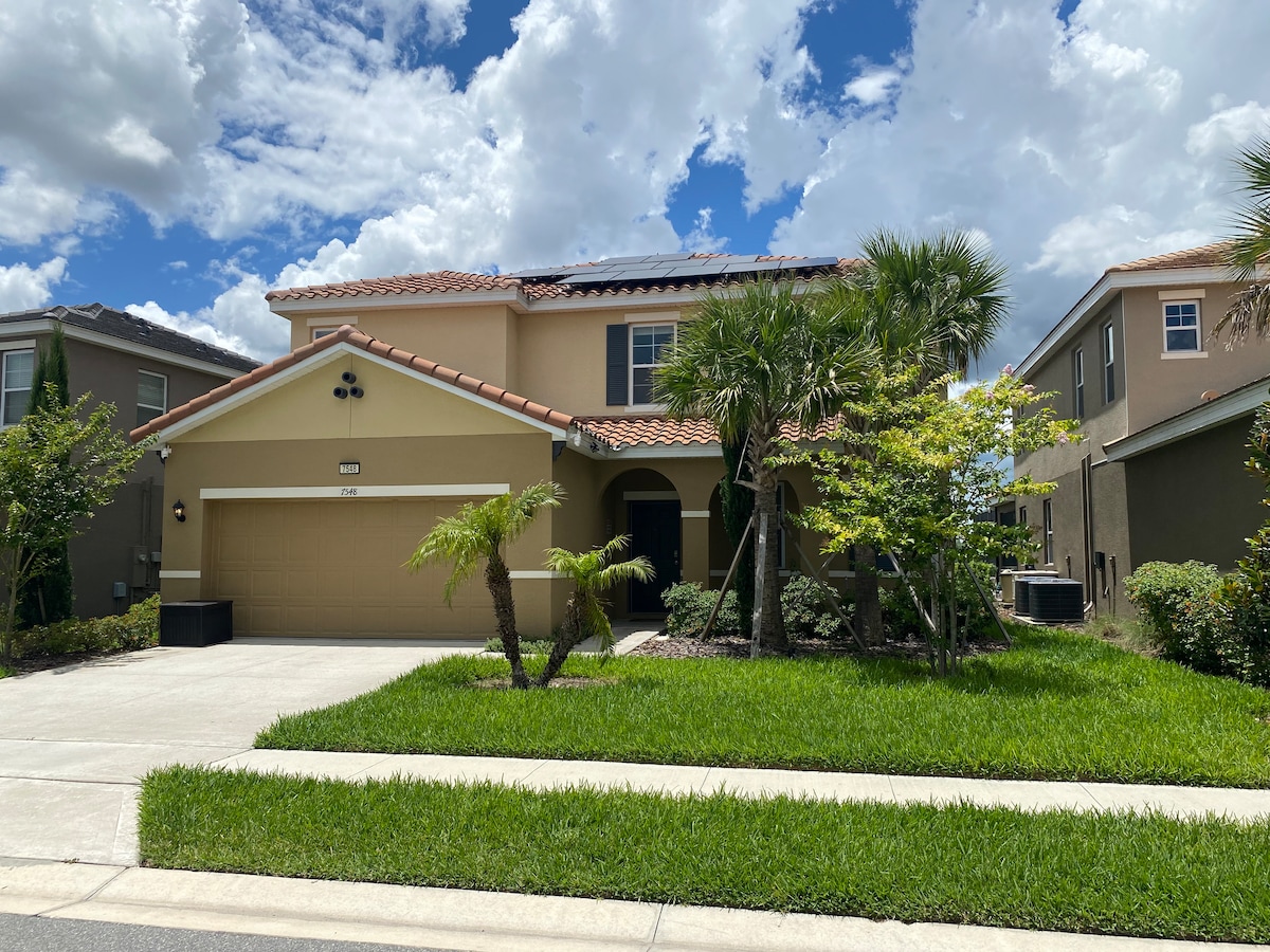 The two-story villa features a warm, beige exterior with a tiled roof and attached garage. Lush green landscaping, including palm and shrubbery, surrounds the entryway, while a paved driveway offers easy access. The bright blue sky with scattered clouds enhances the welcoming atmosphere.
