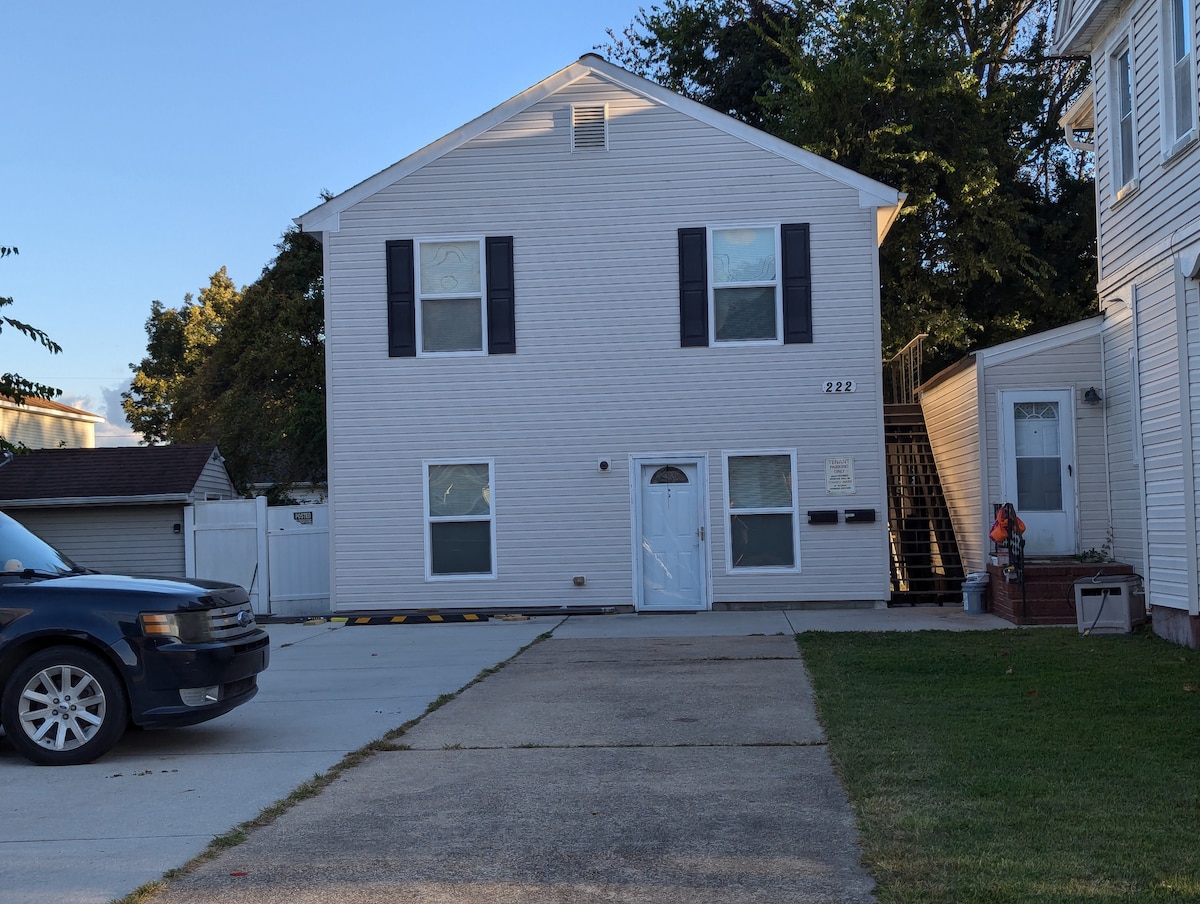 A two-story, light-colored exterior of a house is visible, featuring a white door with a small window and black shutters on each side. A driveway runs alongside the house, leading to off-street parking. Nearby, a set of exterior stairs provides access to a separate entrance.