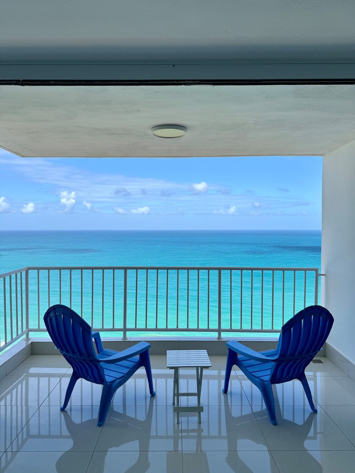 Two blue chairs are positioned on a balcony, facing the ocean. A small white table is placed between the chairs, overlooking the tranquil turquoise waters and clear sky. The scene captures a serene spot for relaxation with an unobstructed coastal view.