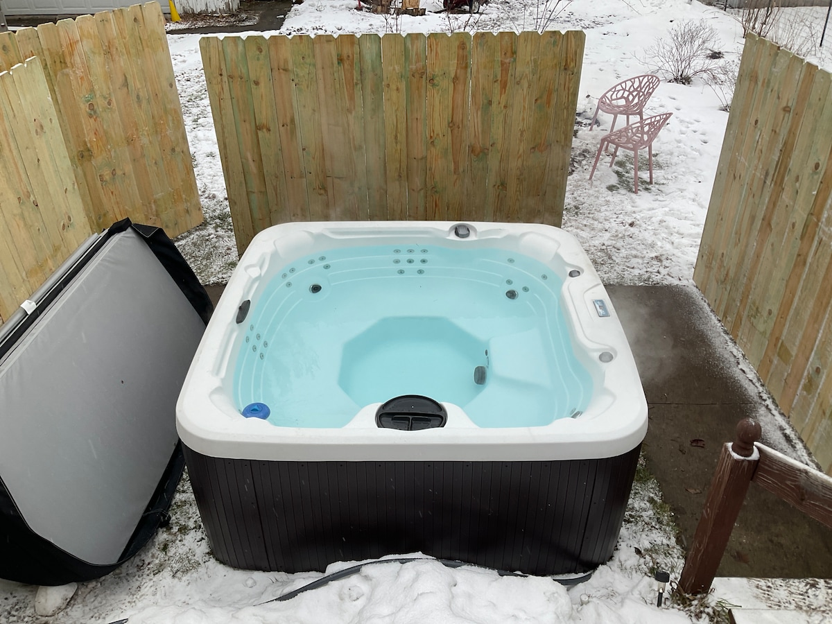 A hot tub is situated on a back deck, surrounded by wooden fencing for privacy. The water is a clear blue, and snow lightly covers the ground nearby. A pink chair is visible in the background, adding a touch of color to the winter scene.