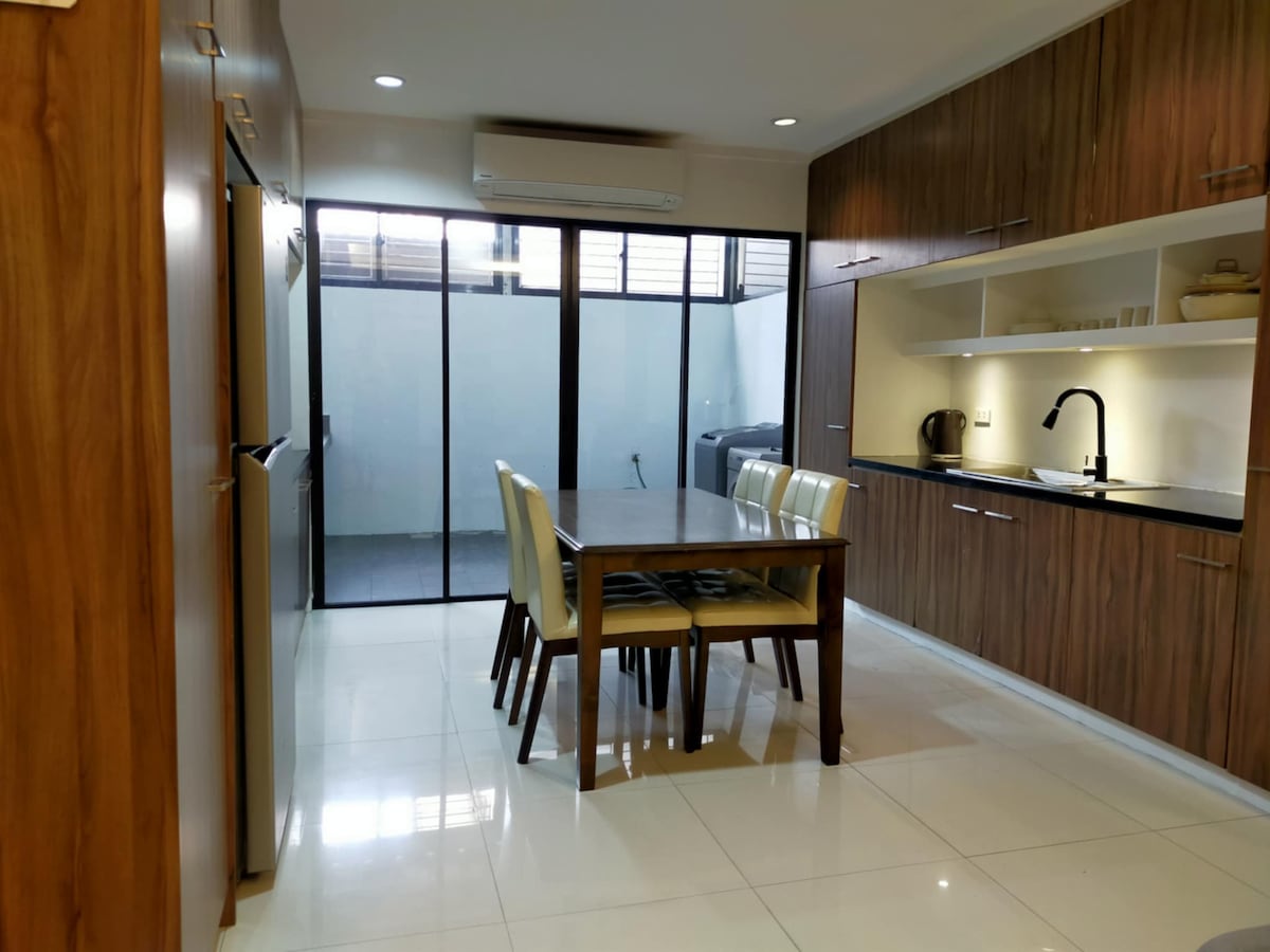 A modern kitchen and dining area feature warm wooden cabinetry and a dining table surrounded by four chairs. Large glass doors allow natural light and lead to a small outdoor space. Appliances such as a kettle and a rice cooker are visible on the countertop.