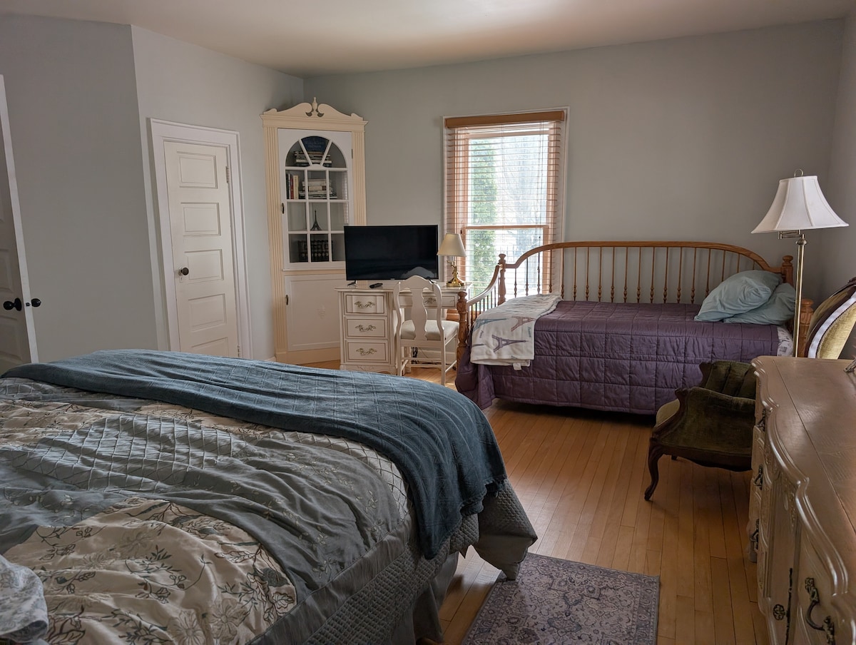 A spacious bedroom is furnished with a king bed draped in a textured quilt. A daybed is positioned nearby, alongside a wooden desk and chair. Natural light filters through the window, complemented by a tall lamp. A white cabinet with glass doors adds character to the room.