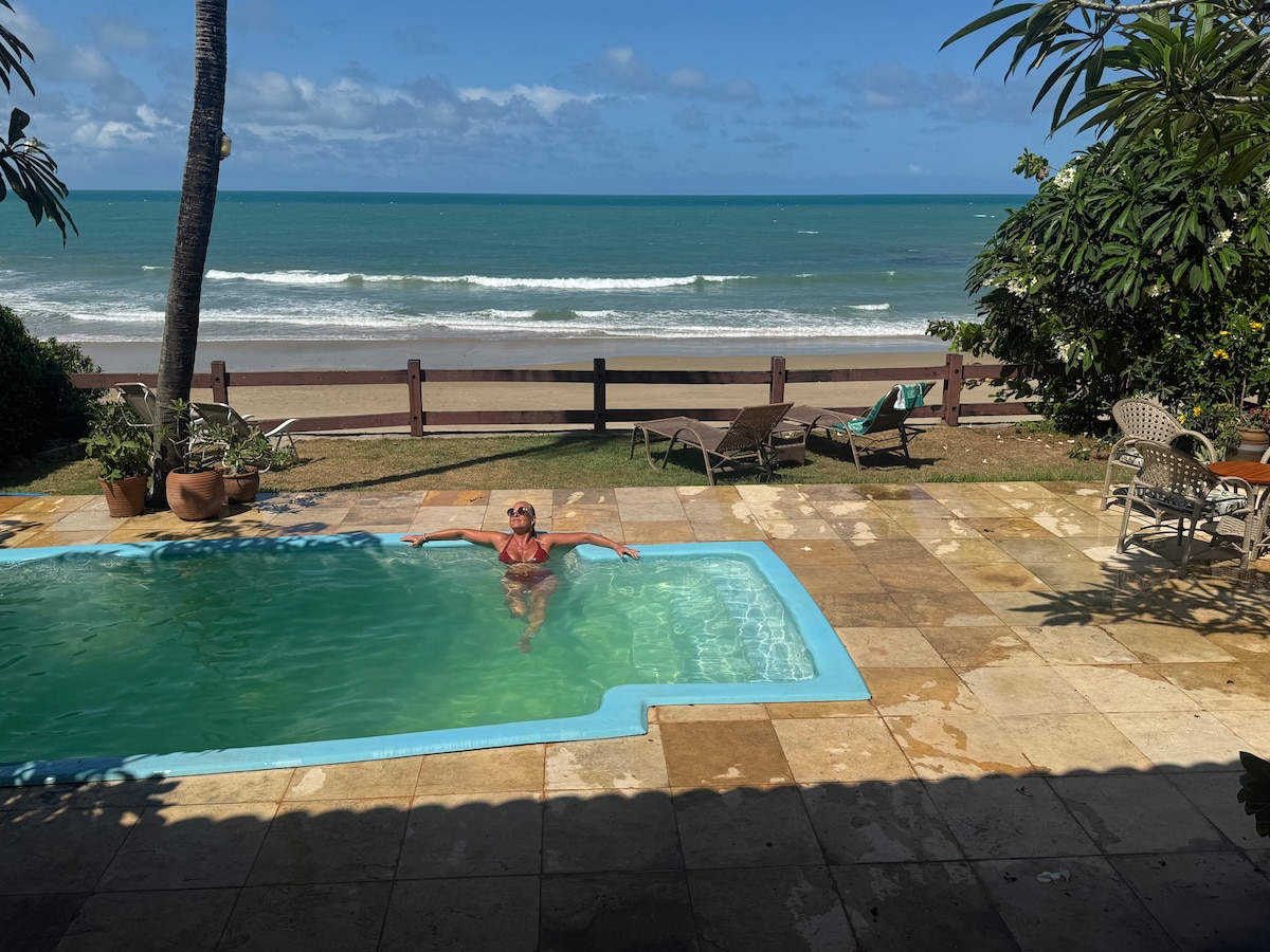 A private swimming pool is situated in a stone-tiled outdoor area. Sun loungers are positioned nearby, with lush greenery framing the space. A view of the beach and ocean is visible beyond the pool, with gentle waves lapping against the shore under a clear blue sky.