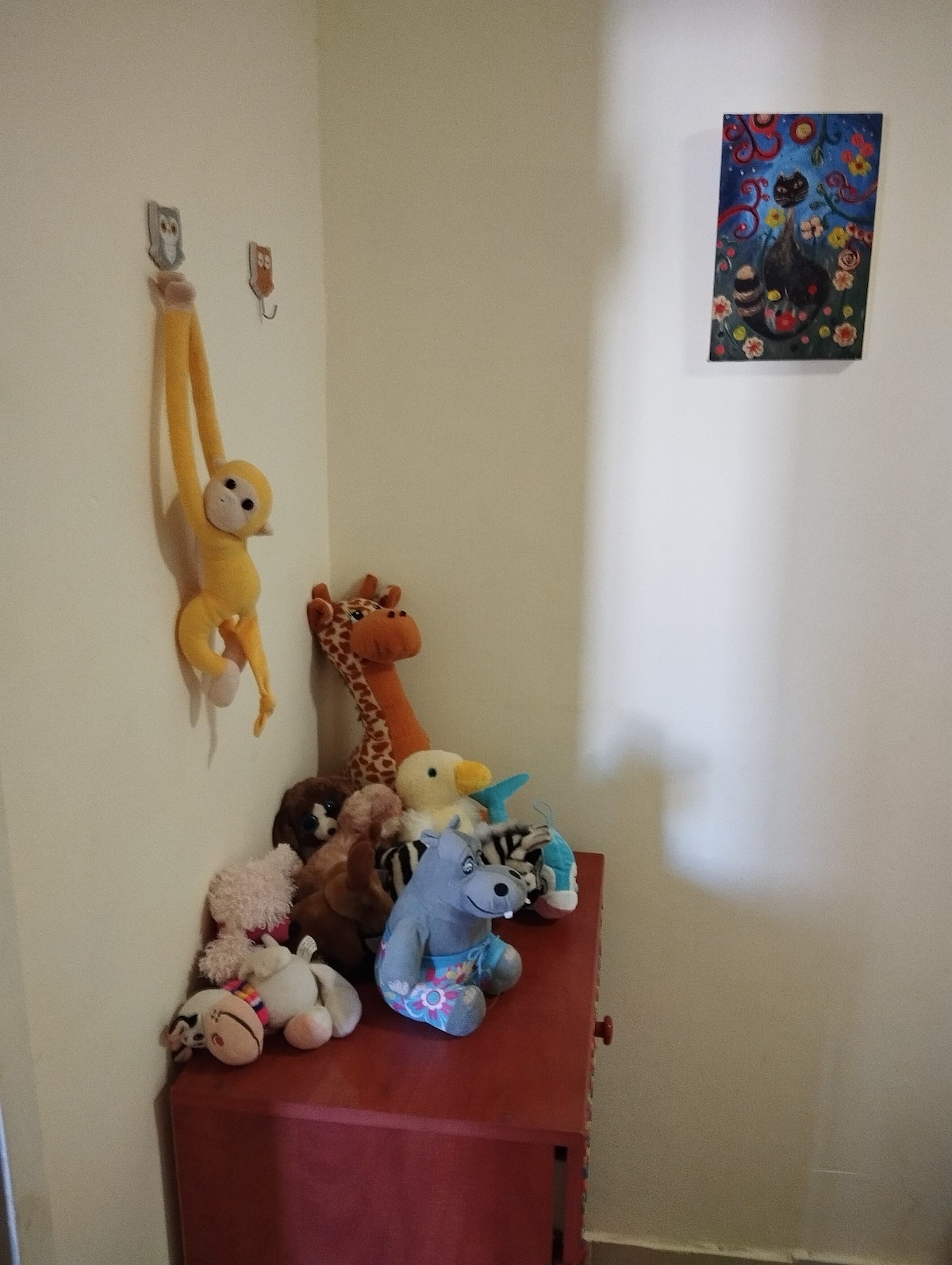A cozy corner displays a colorful assortment of stuffed animals, including a monkey, giraffe, and unicorn, arranged around a wooden chest. A vibrant artwork featuring cats adorns the wall, while natural light filters in through a nearby window.