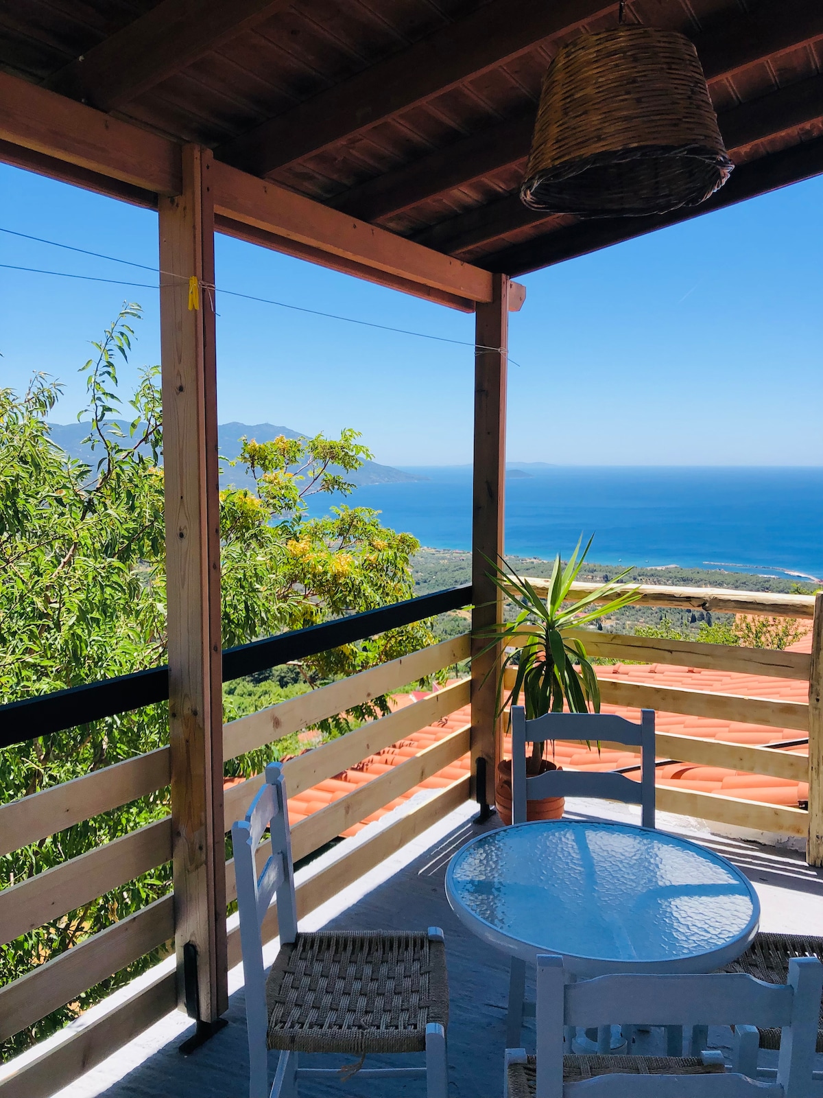 An outdoor balcony offers a clear view of the sea and surrounding landscape. A round glass table is set with two white chairs, while a potted plant adds a touch of greenery. The wooden railing provides a rustic charm, enhancing the sense of openness to the scenic backdrop.