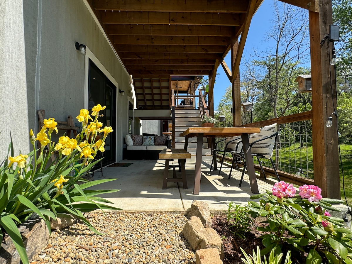 An outdoor patio space is visible, featuring a wooden table surrounded by chairs. Colorful flowers, including yellow and pink blooms, are positioned at the edges. A stone pathway leads to the area, with a glimpse of stairs and the backyard beyond.
