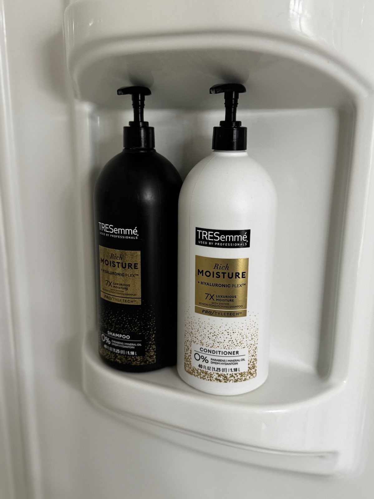 A shower caddy features two large bottles of TRESemmé hair care products. The black bottle is labeled as shampoo, while the white bottle is marked as conditioner. Both bottles are mounted against a smooth, white wall, with clearly visible pump tops for easy use.