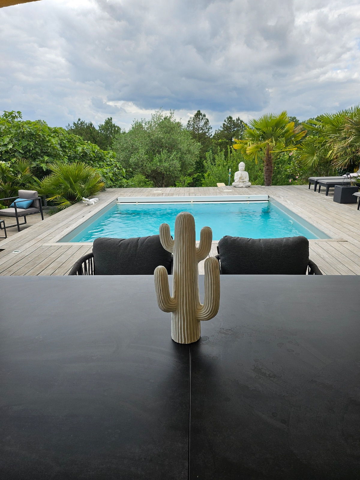 A cactus centerpiece is displayed on a dark table, with a clear view of a swimming pool surrounded by wooden decking and lush greenery. A soothing statue is positioned near the pool, creating a serene outdoor setting.