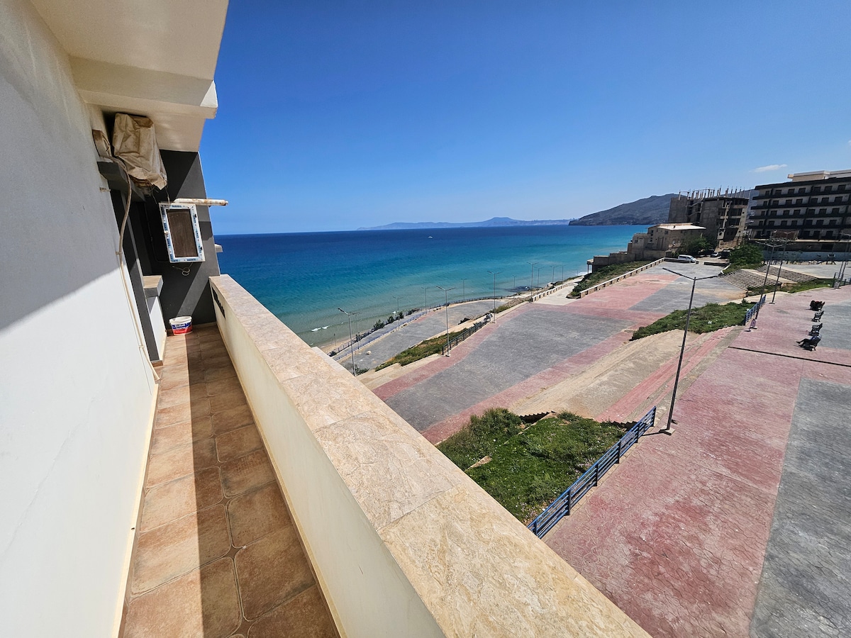 A spacious balcony provides a clear view of the sandy beach and turquoise water stretching into the horizon. The scene is complemented by a serene sky, enhancing the coastal atmosphere. The area features well-maintained pathways and green spaces, suitable for relaxation and outdoor activities.