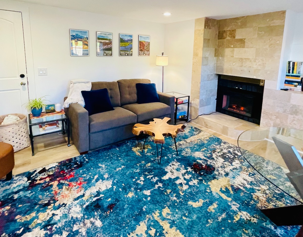 A cozy seating area features a comfortable sofa accented with navy blue pillows. A coffee table made of natural wood sits atop a vibrant area rug with abstract patterns. Warm light is provided by a nearby lamp, and a stylish electric fireplace enhances the space.