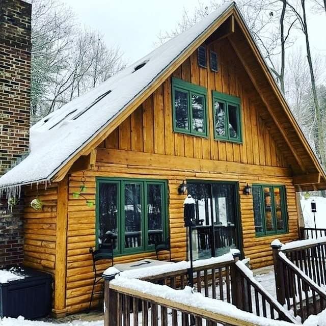 The exterior of a cozy wooden cabin is shown, adorned with snow on the roof and surrounding areas. Green-framed windows complement the natural wood finish. A welcoming deck with railings is visible, complete with chairs, creating an inviting space for relaxation amidst the winter setting.
