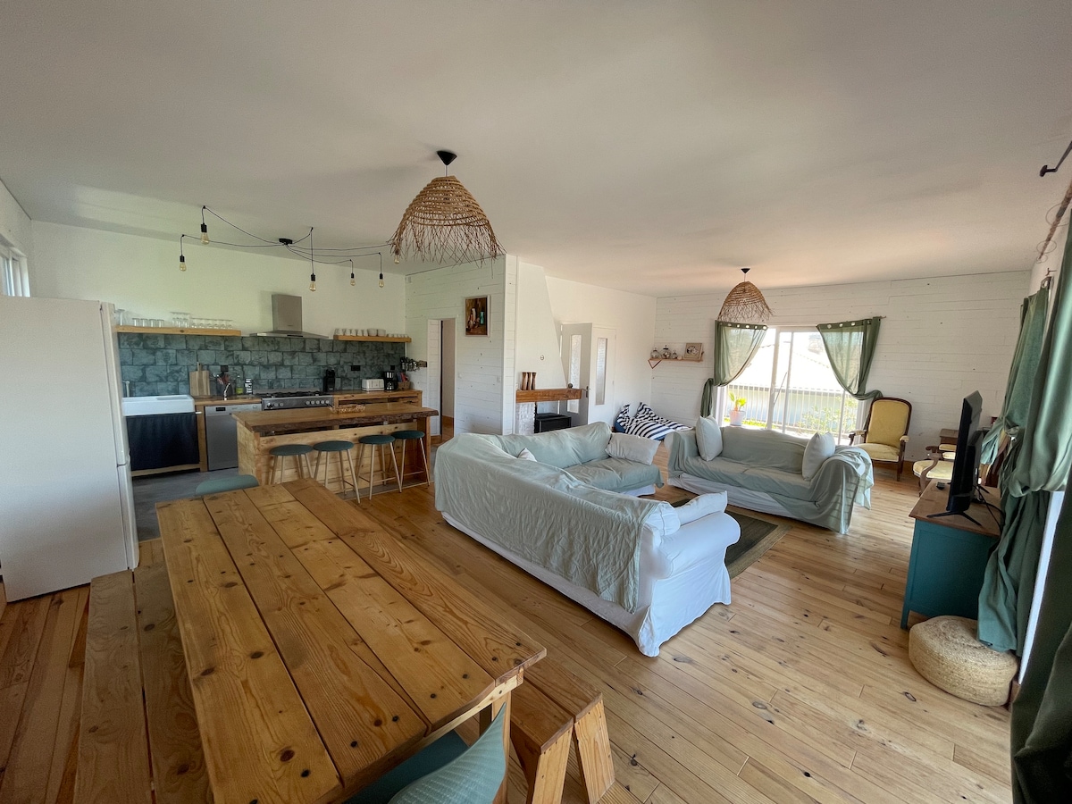 A spacious living area is designed with natural wood accents and features several light-colored sofas arranged for relaxation. A large wooden dining table sits prominently, alongside a well-equipped kitchen with a stone backsplash. Large windows allow ample natural light, enhancing the overall bright atmosphere.