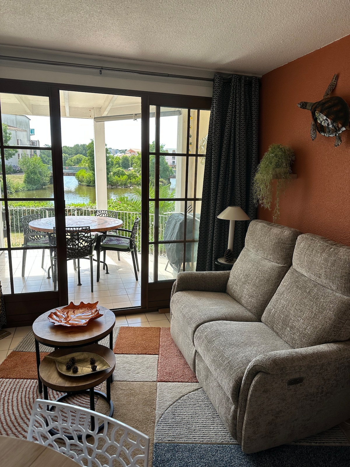 A living area is shown with a plush sofa facing large glass doors that open to a terrace overlooking greenery and a lake. A small round table is set beside the sofa, with decorative items on it. A lamp and wall decor enhance the space.