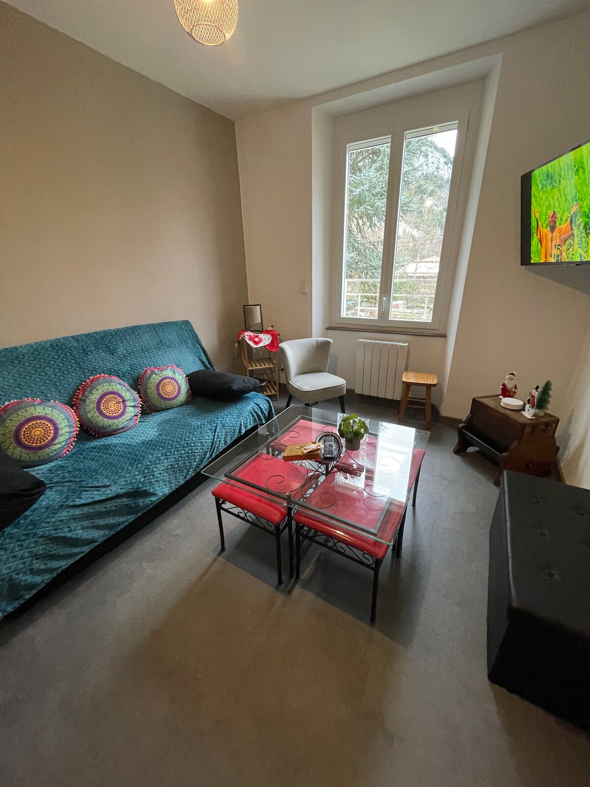 A living area features a teal sofa bed adorned with decorative cushions. A glass coffee table sits at the center, surrounded by a cozy armchair and a wooden stool. Natural light streams in through a large window, illuminating the space.