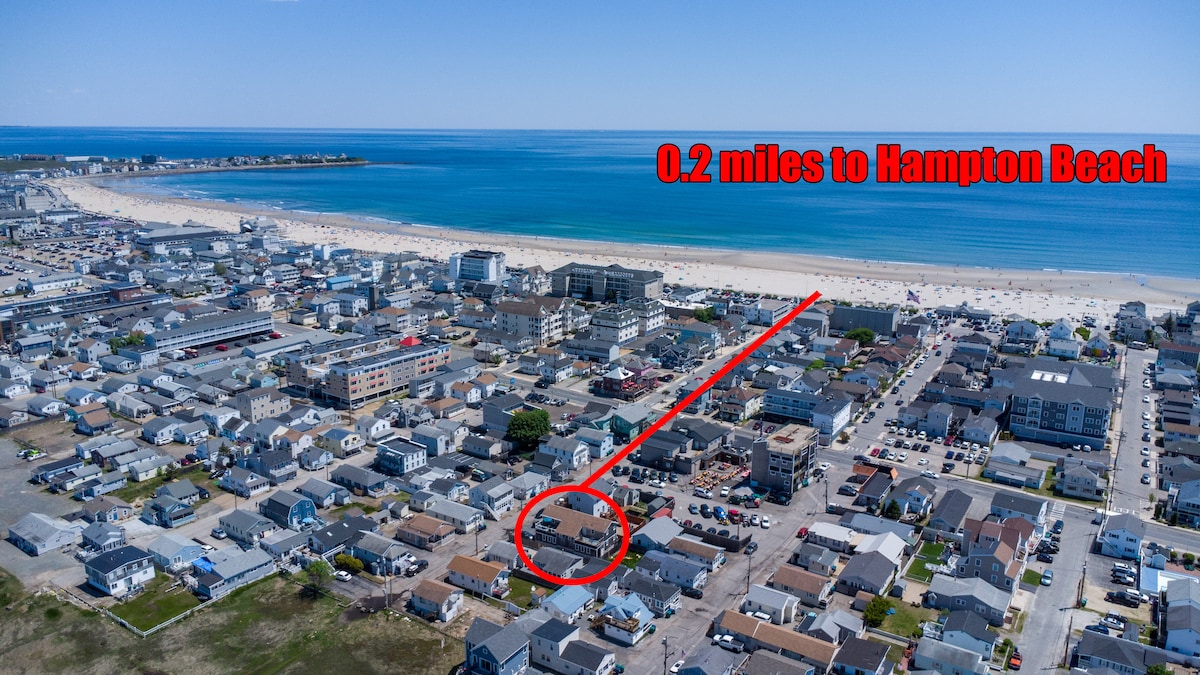 An aerial view captures the proximity to Hampton Beach, indicated by a red arrow, featuring sandy shores and blue ocean waters. Residential structures and recreational areas surrounding the beach are visible, emphasizing the close distance of 0.2 miles to the shoreline.