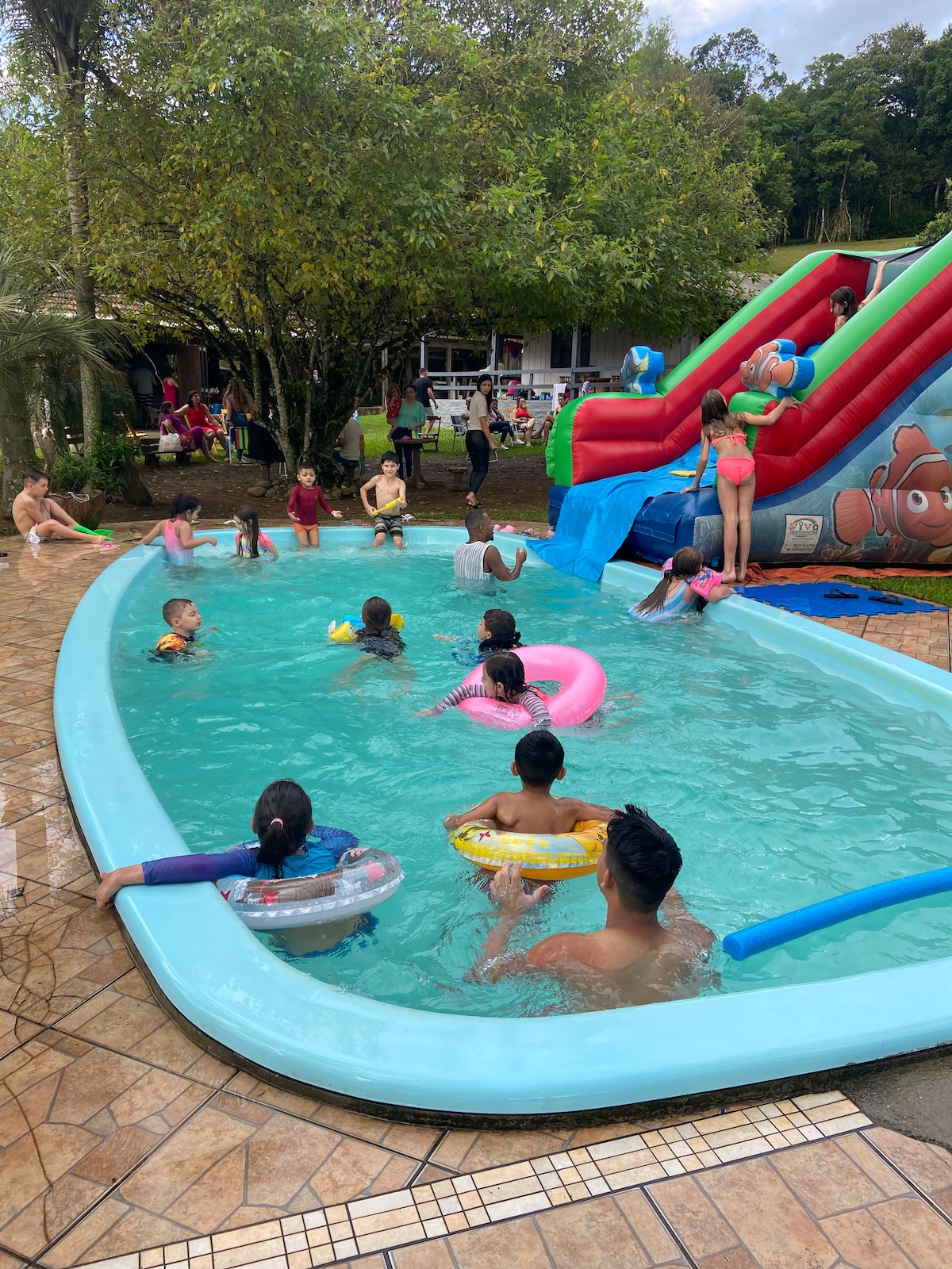 A vibrant outdoor pool area is filled with children enjoying themselves, floating on colorful inflatable rings. A water slide adds excitement nearby. Lush greenery surrounds the scene, complementing the cheerful atmosphere of the gathering.