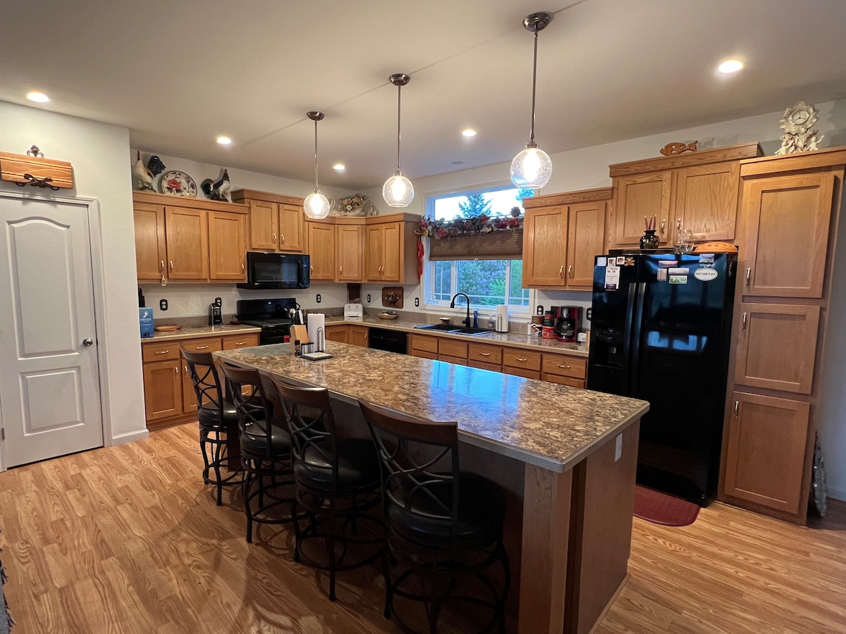 The kitchen features wooden cabinetry, a large island with seating for four, and modern appliances, including a black refrigerator and stovetop. Warm lighting is provided by pendant fixtures, and a window offers natural light, enhancing the space's functionality.