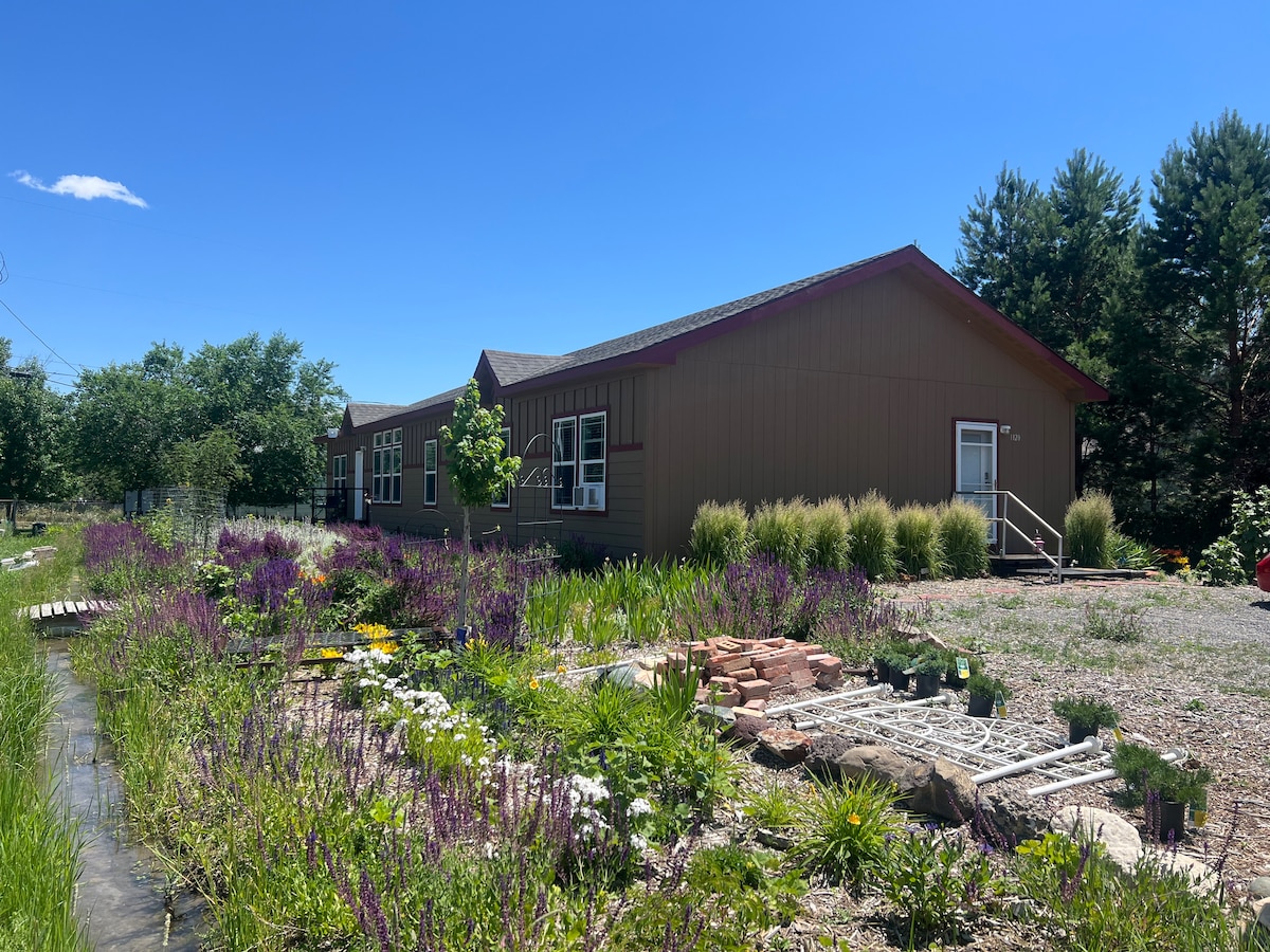 A spacious building is viewed from the side, surrounded by green grass and colorful wildflowers. A small stream runs along the property, bordered by natural stones. The clear blue sky is visible above, accentuating the peaceful outdoor setting.