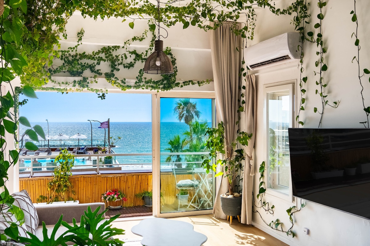The living room features wall-to-wall windows opening towards the sea, with views of the harbor and Gordon Beach. Natural plants adorn the space, complemented by a comfortable sofa and a wall-mounted television. Soft curtains frame the windows, allowing for light control.