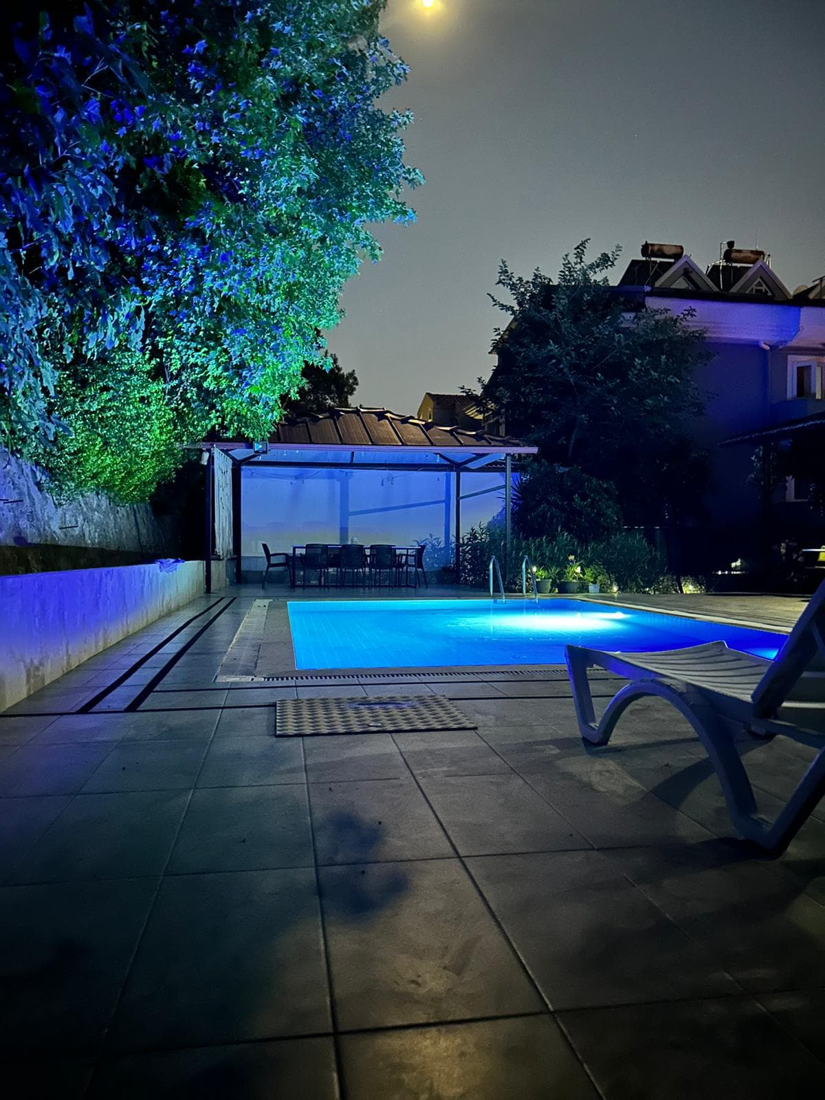A tranquil outdoor area features a pool illuminated in soft blue light, surrounded by lush greenery. A covered seating area is visible in the background, with a partially shaded patio that complements the serene environment.