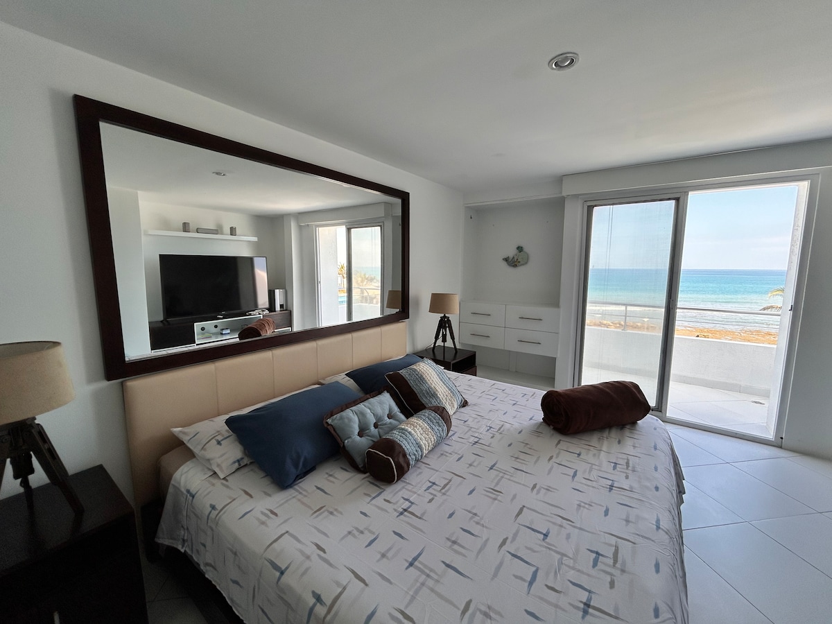 A spacious bedroom features a large bed adorned with a patterned bedspread and decorative pillows. Natural light fills the room through glass sliders leading to a serene ocean view. A flat-screen television is mounted on a wall opposite the bed, complemented by sleek nightstands.