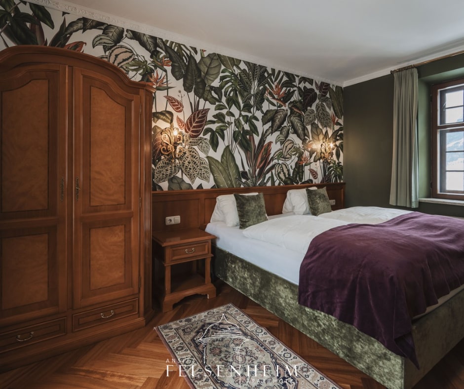 A cozy bedroom features a spacious bed with a plush maroon blanket. Dark green walls are complemented by a vibrant botanical wallpaper. A wooden armoire provides storage, while a bedside table rests next to the bed. Natural light enters through a window with wooden accents.
