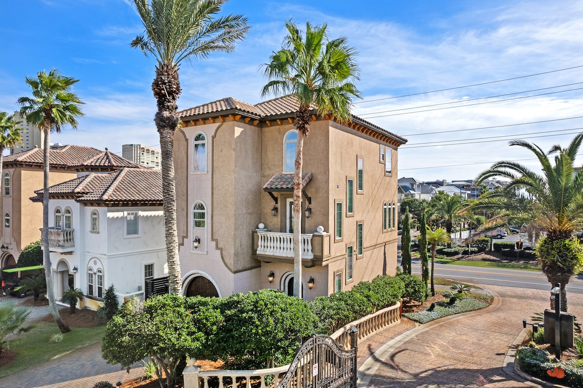 The exterior of the Mediterranean home is displayed, featuring stucco walls and ornate architectural details. Lush palm trees and manicured landscaping frame the property, while a scenic road runs alongside. The inviting entrance showcases a balcony with decorative elements, enhancing the overall elegance of the design.