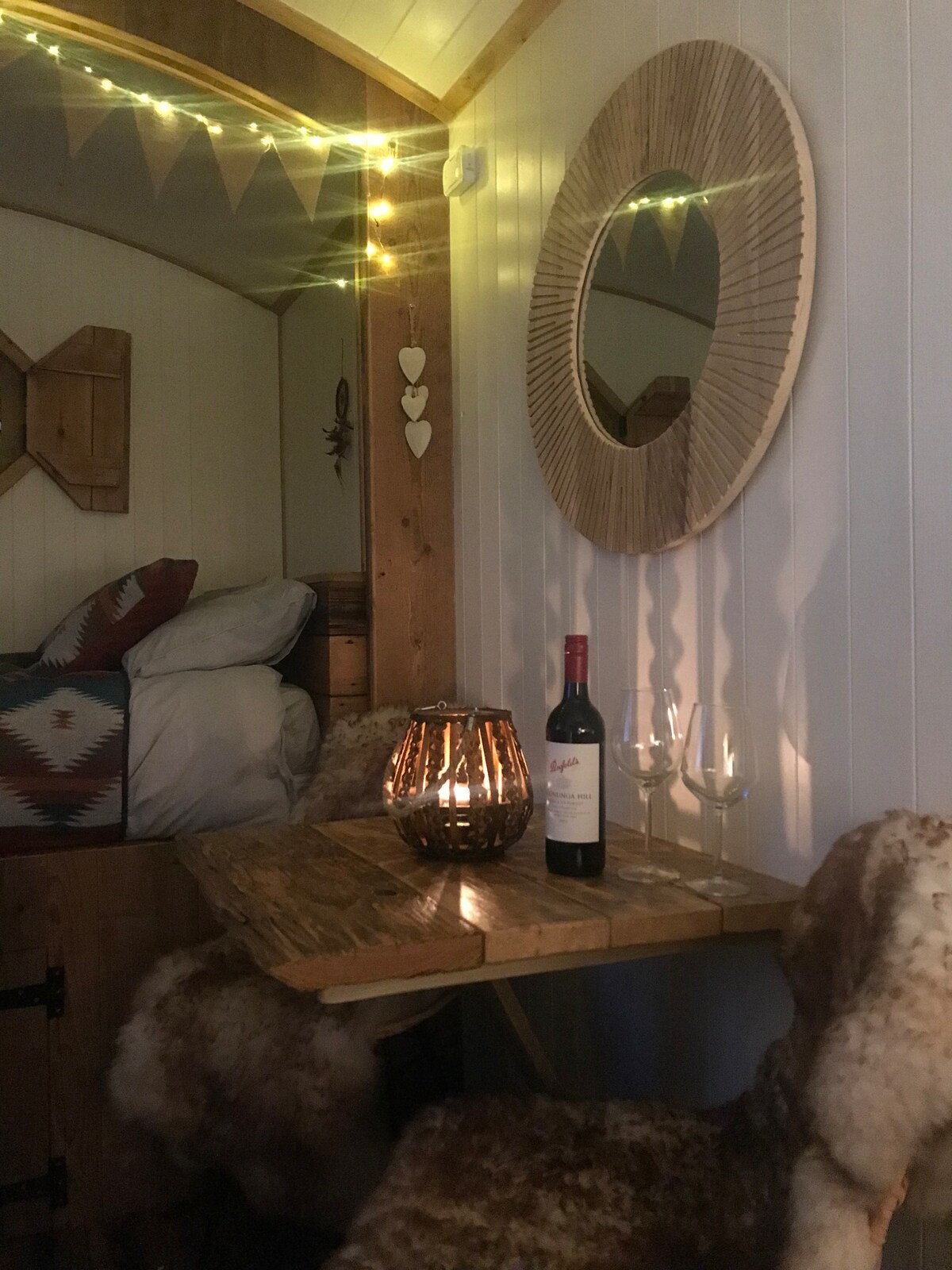 A cozy seating area is illuminated by soft string lights. A round mirror hangs on the wall above a rustic table, which holds a bottle of wine and two glasses. Textured chairs covered in soft fur add warmth.