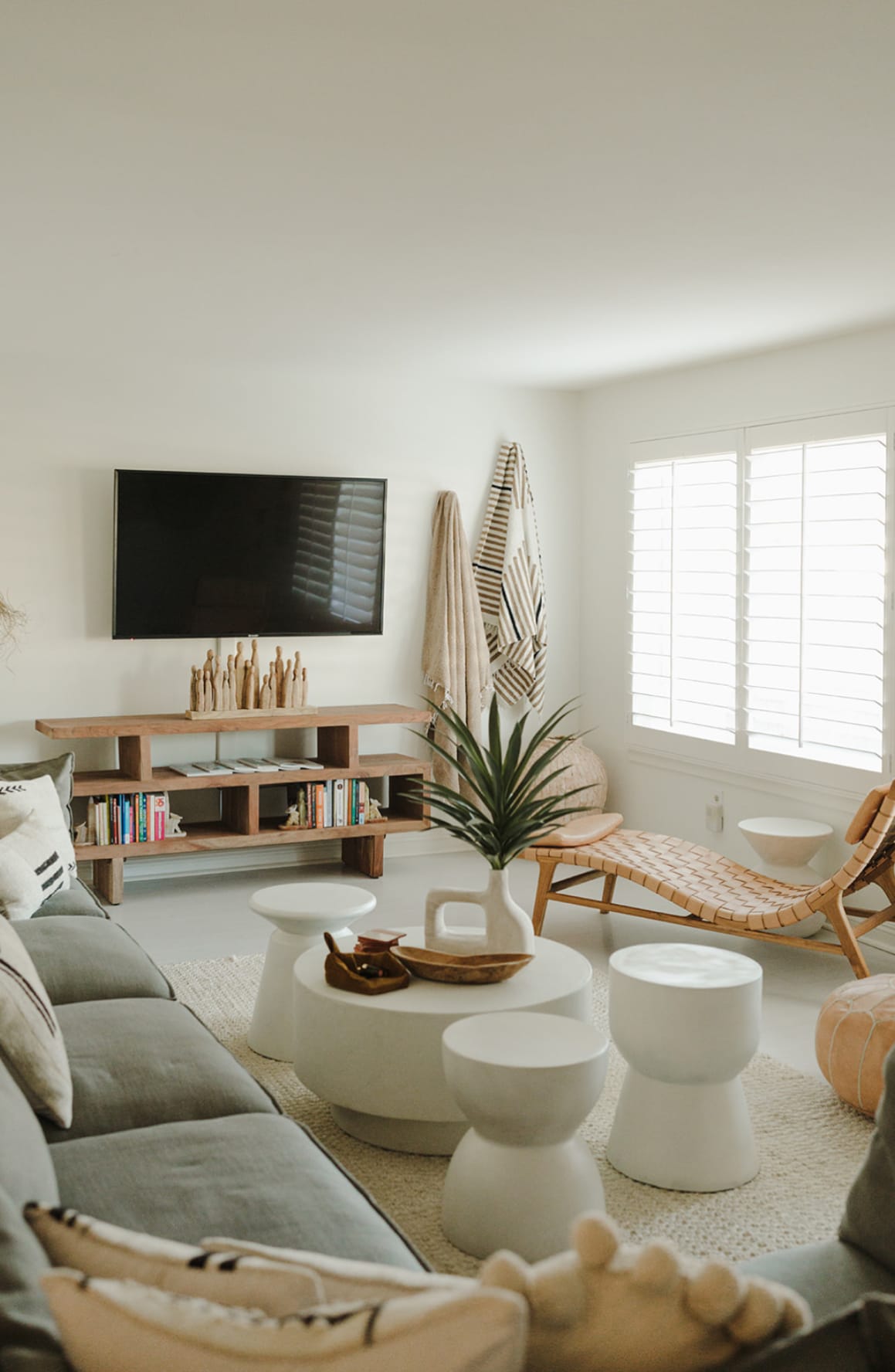 A modern living area features a comfortable grey sofa with decorative cushions, complemented by a series of white side tables. A wooden shelf displays books and decorative items, while a flat-screen TV is mounted on the wall. Natural light filters through the window with plantation shutters.
